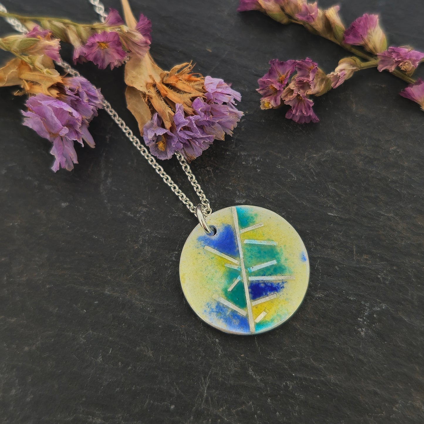 A round silver pendant with abstract silver lines and blue and turquoise enamels blending into pale yellow. Suspended from a silver chain. Small. Pictured on slate with flowers.