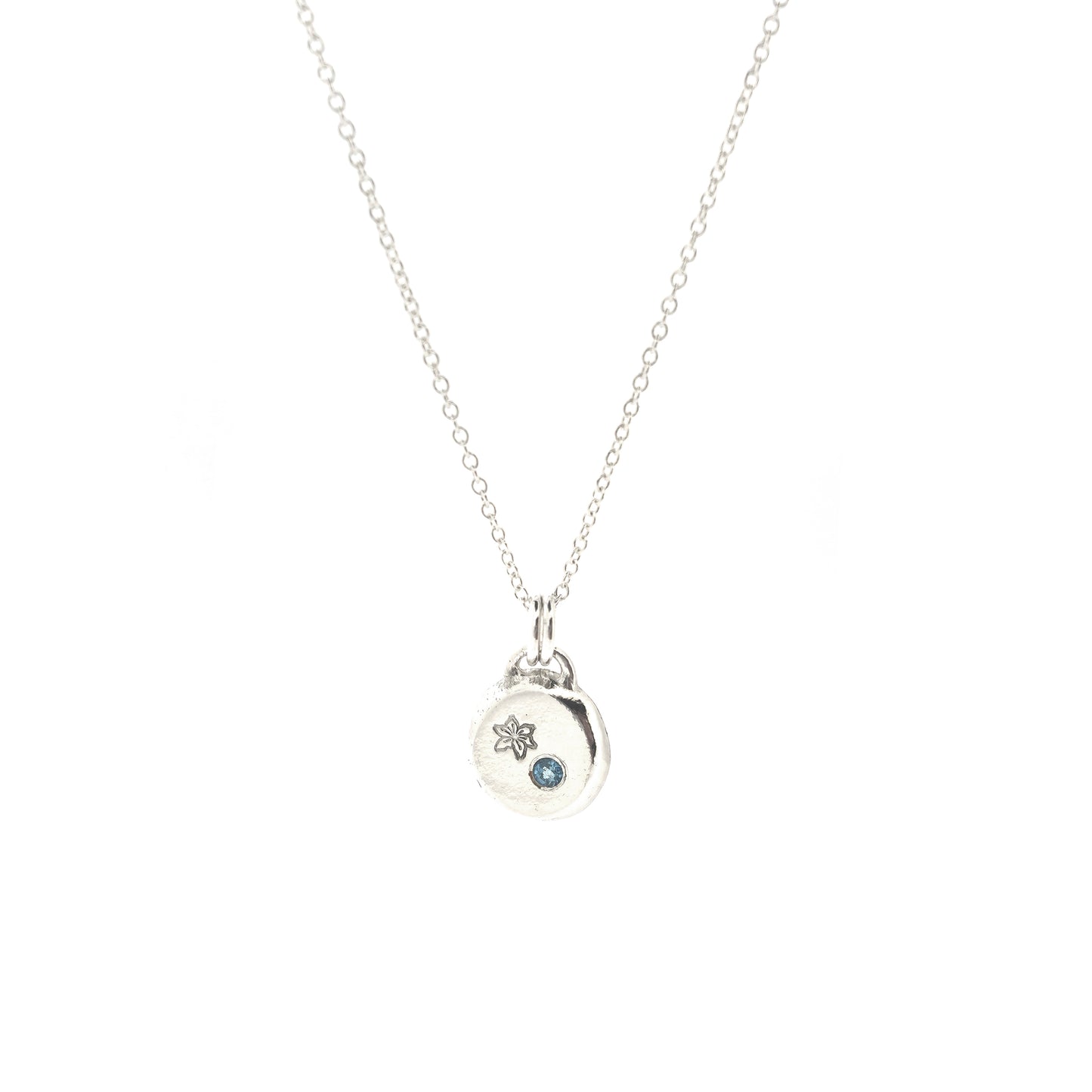 A silver round flat pebble pendant set with a round blue topaz gemstone (December birthstone) and engraved with a poinsettia flower (December birth flower) on a silver chain.