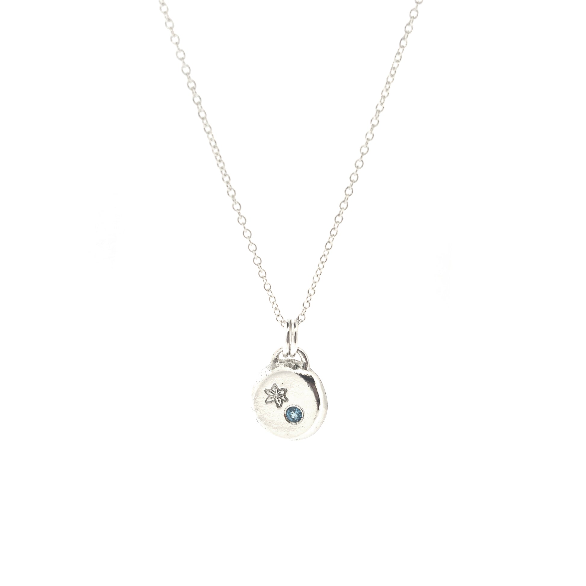 A silver round flat pebble pendant set with a round blue topaz gemstone (December birthstone) and engraved with a poinsettia flower (December birth flower) on a silver chain.