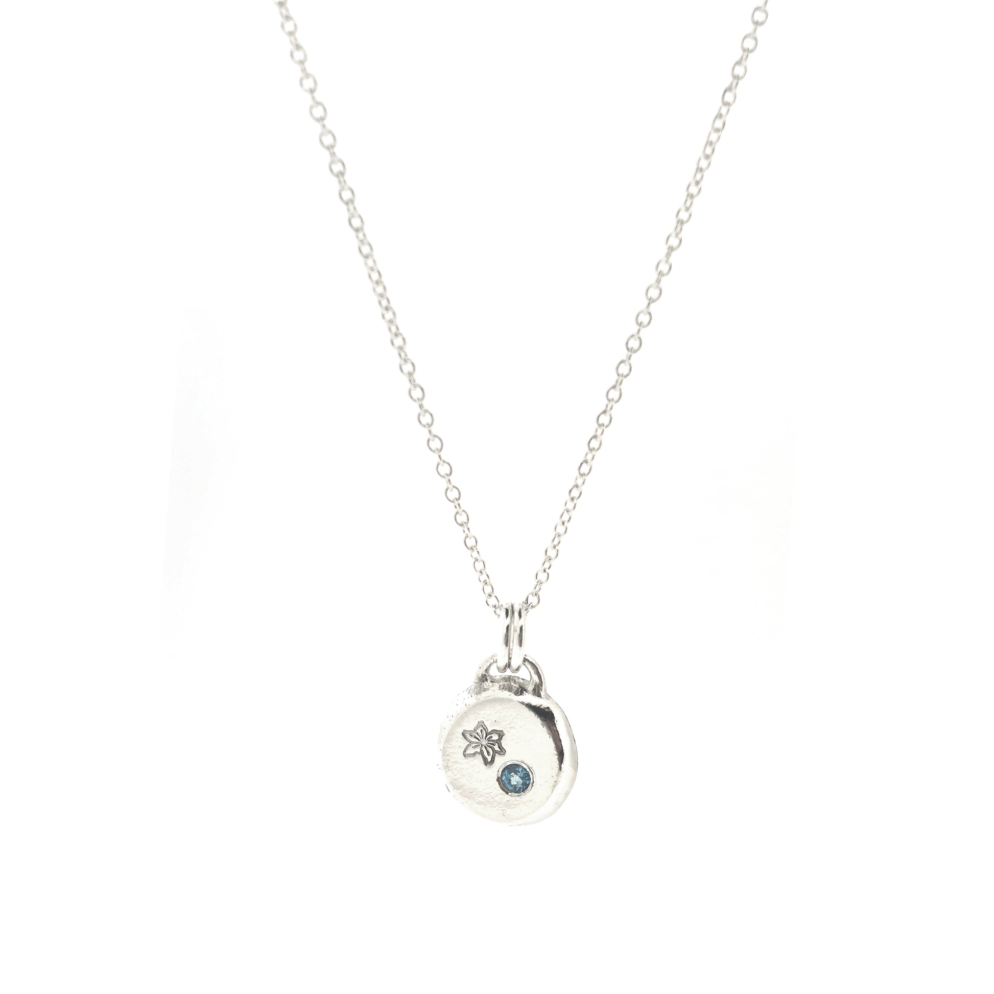 A silver round flat pebble pendant set with a round blue topaz gemstone (December birthstone) and engraved with a poinsettia flower (December birth flower) on a silver chain.