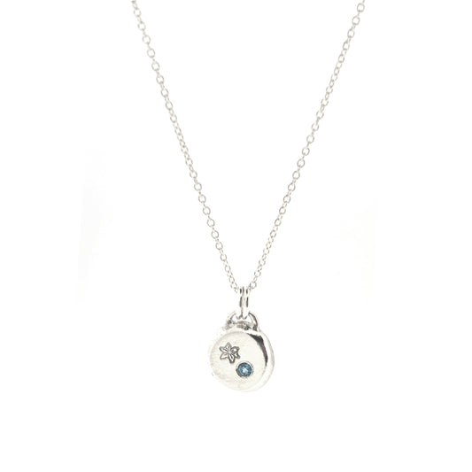 A silver round flat pebble pendant set with a round blue topaz gemstone (December birthstone) and engraved with a poinsettia flower (December birth flower) on a silver chain.