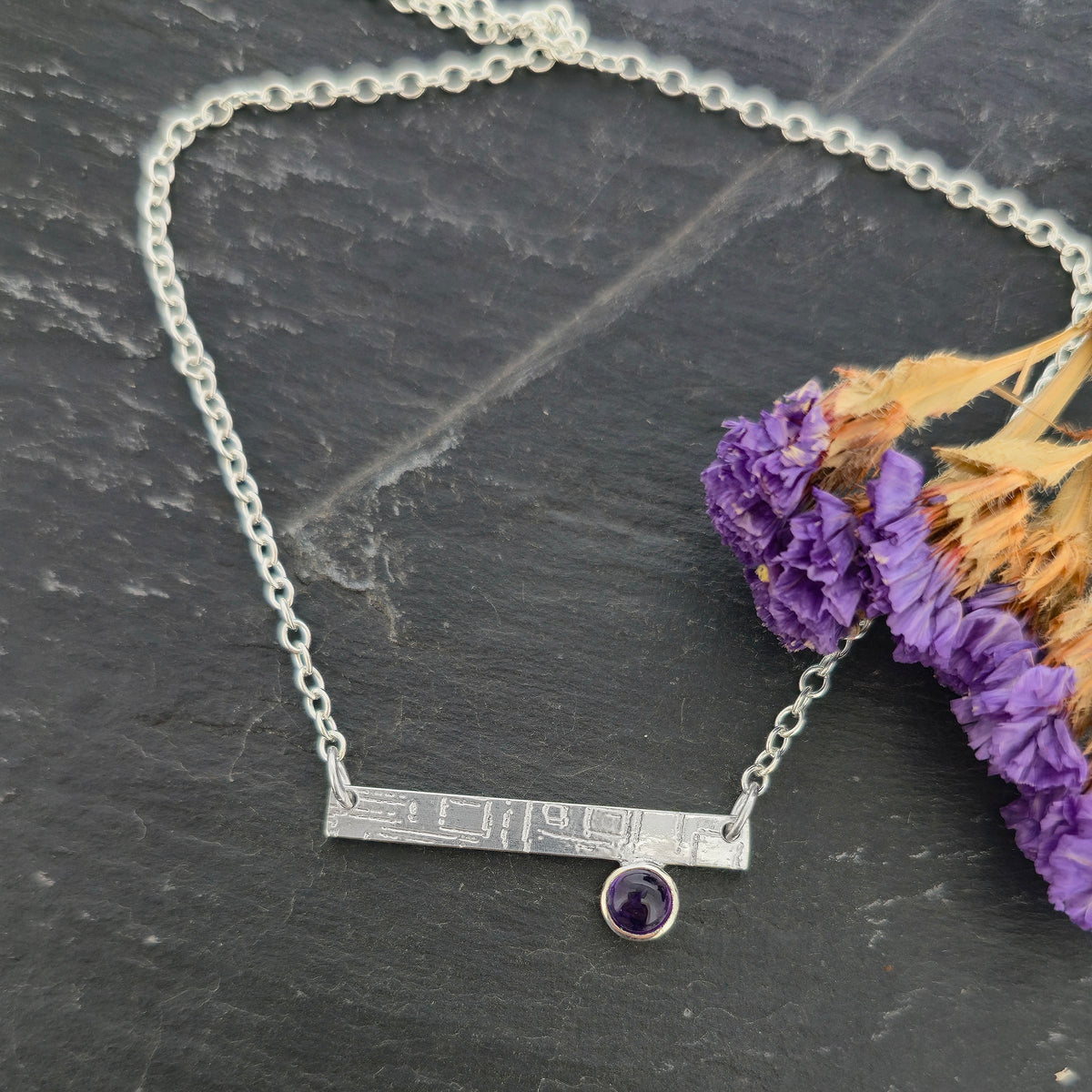 A handmade sterling silver bar necklace with engraved square pattern and a round bezel-set purple amethyst gemstone, displayed on a dark slate surface next to dried purple flowers. Mijoux Jewellery.