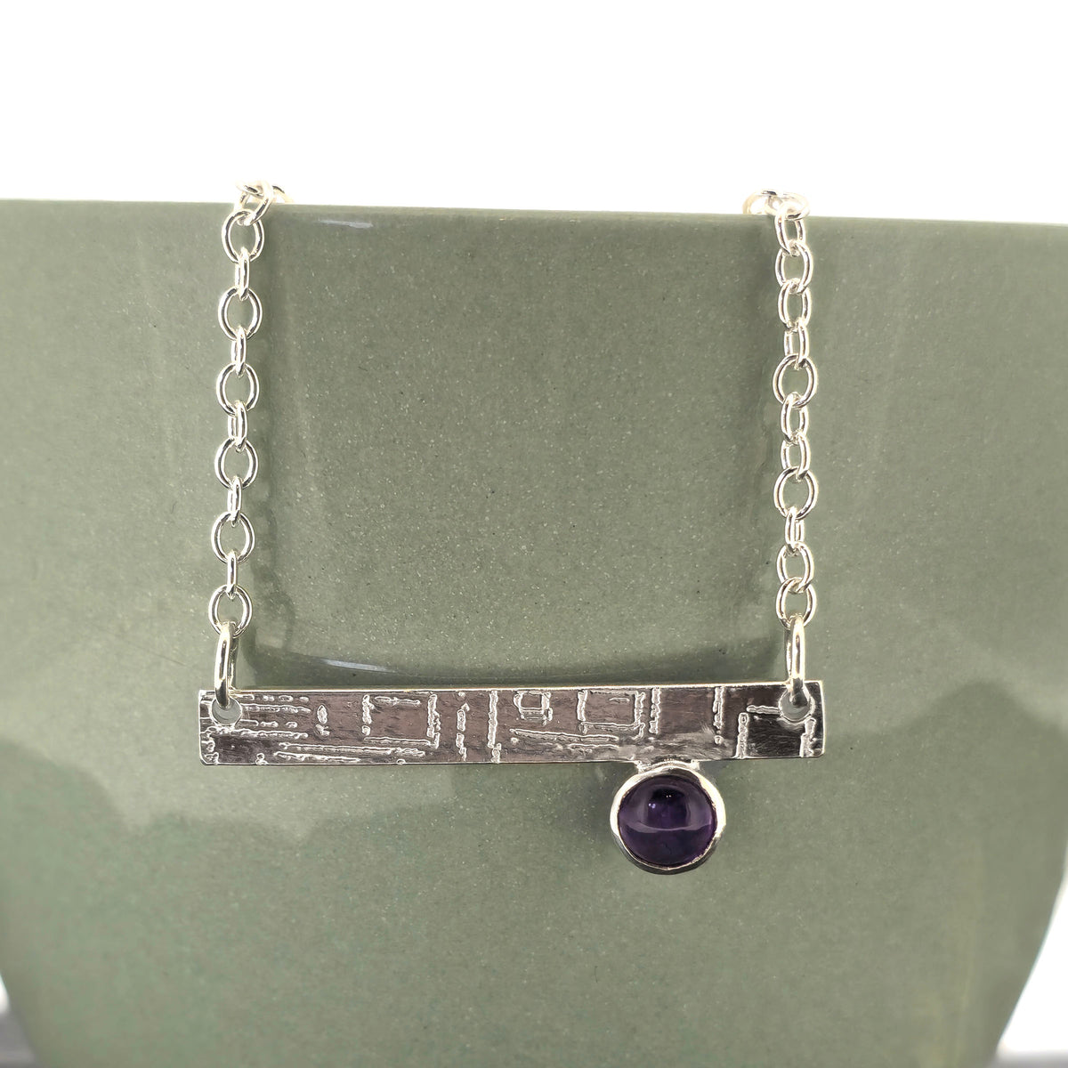 A handmade sterling silver bar necklace with a textured horizontal bar and a small round purple amethyst gemstone, displayed on a matte green ceramic stand. Mijoux Jewellery.