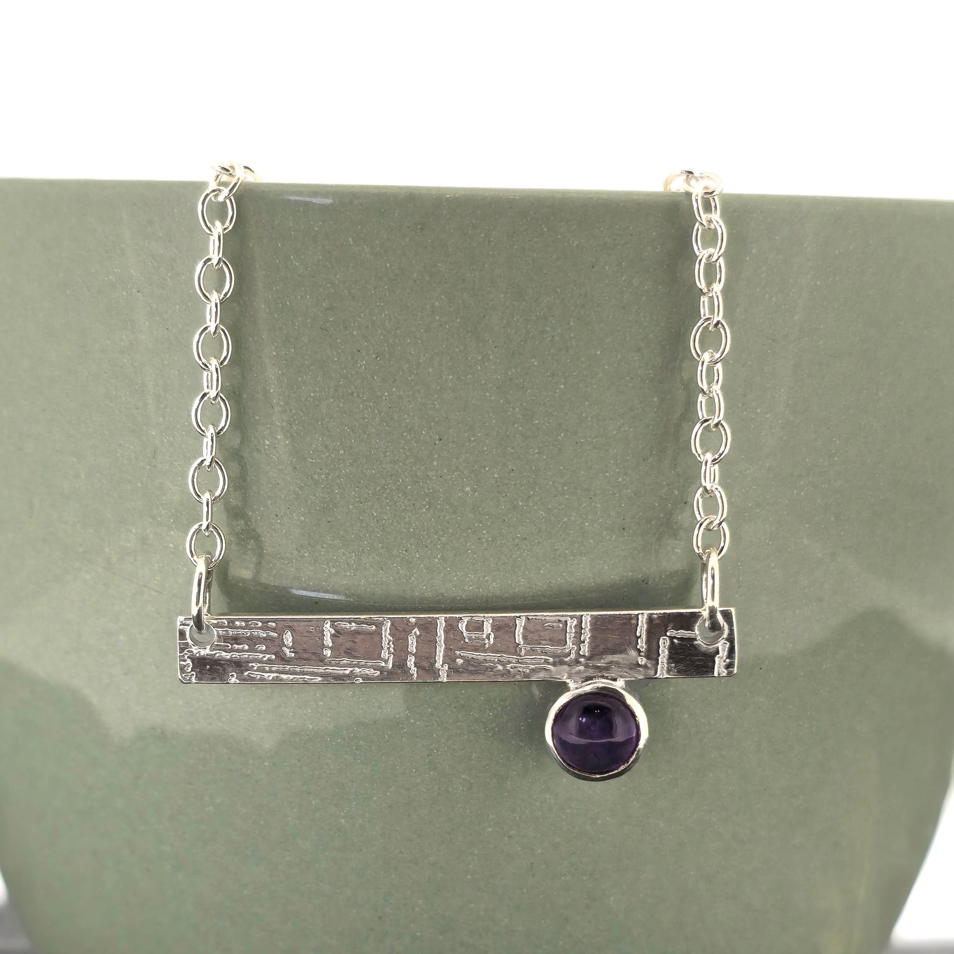 A handmade sterling silver bar necklace with a textured horizontal bar and a small round purple amethyst gemstone, displayed on a matte green ceramic stand. Mijoux Jewellery.