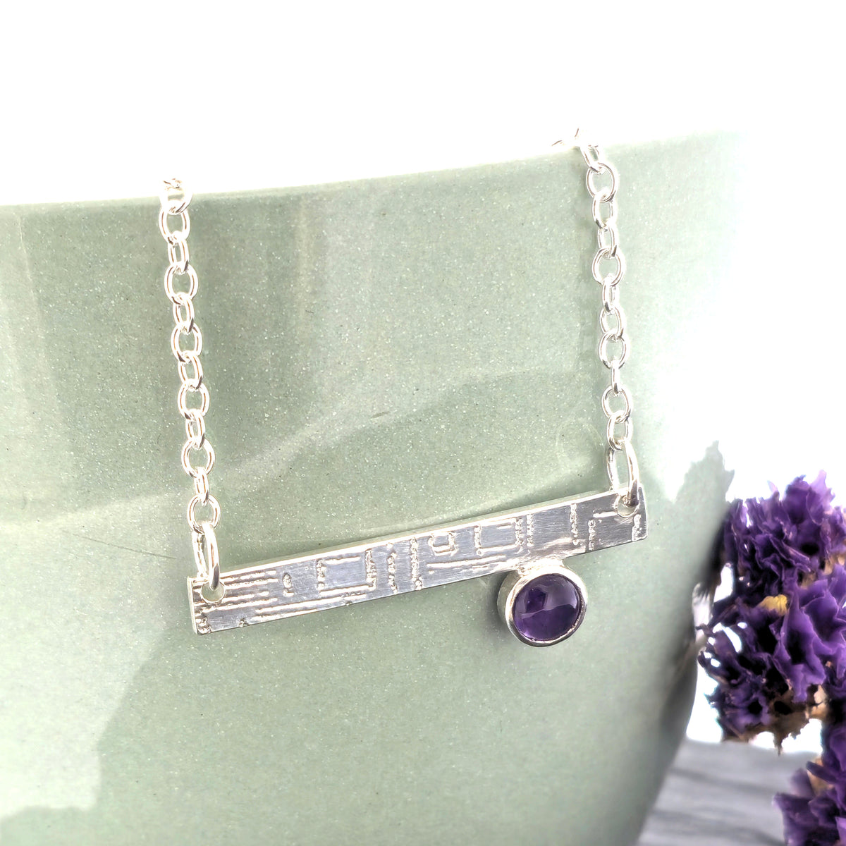 A handmade sterling silver bar necklace with a textured horizontal bar and a small round purple amethyst gemstone, displayed on a matte green ceramic stand with purple flowers. Mijoux Jewellery.