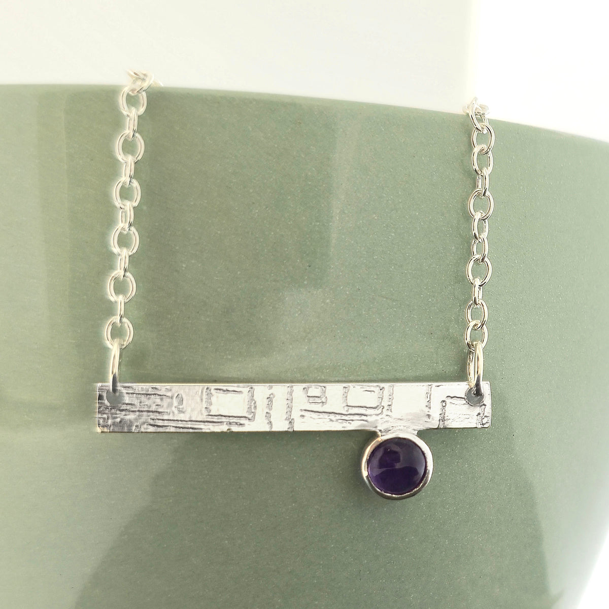 A handmade sterling silver bar necklace with a textured horizontal bar and a small round purple amethyst gemstone, displayed on a matte green ceramic stand. Mijoux Jewellery.