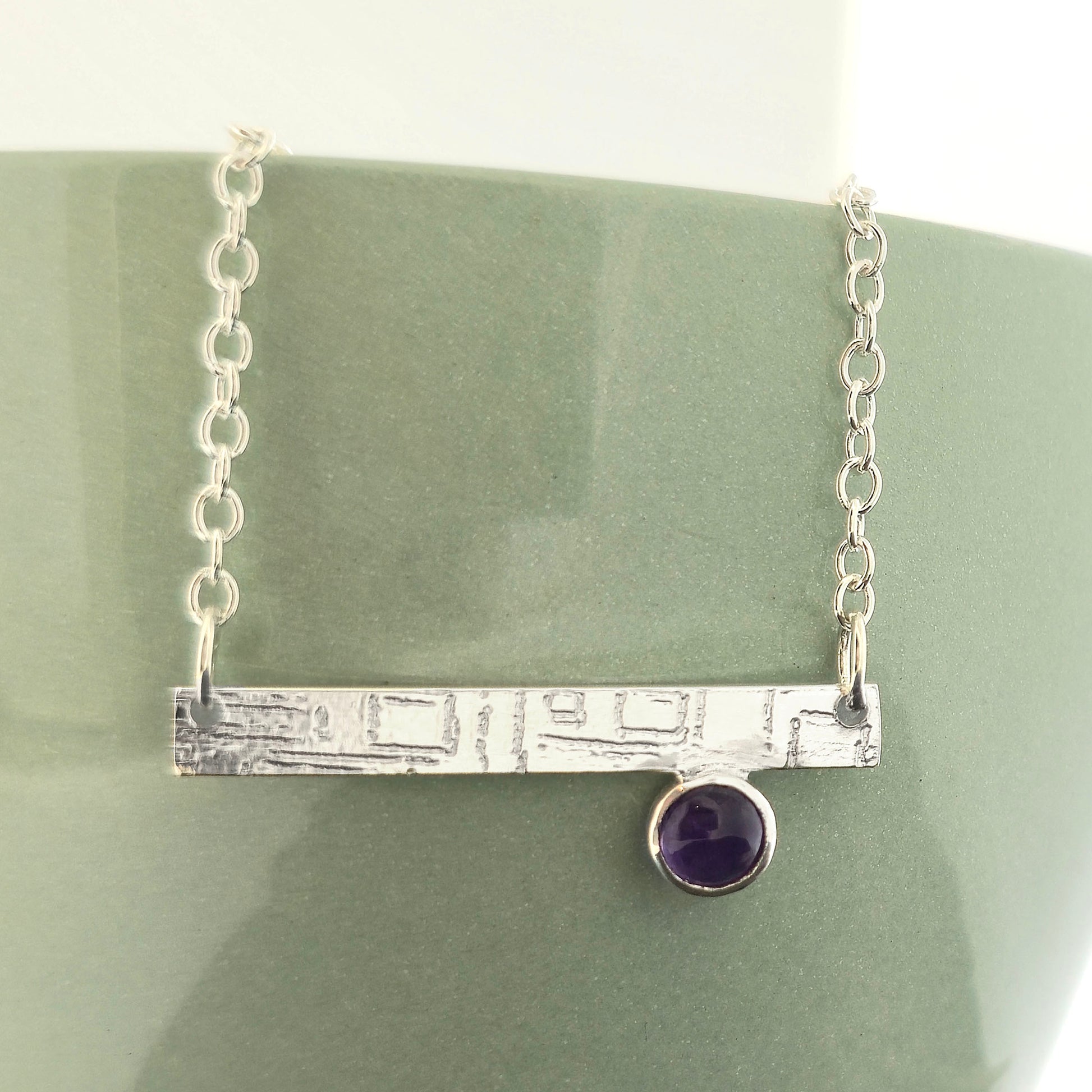 A handmade sterling silver bar necklace with a textured horizontal bar and a small round purple amethyst gemstone, displayed on a matte green ceramic stand. Mijoux Jewellery.