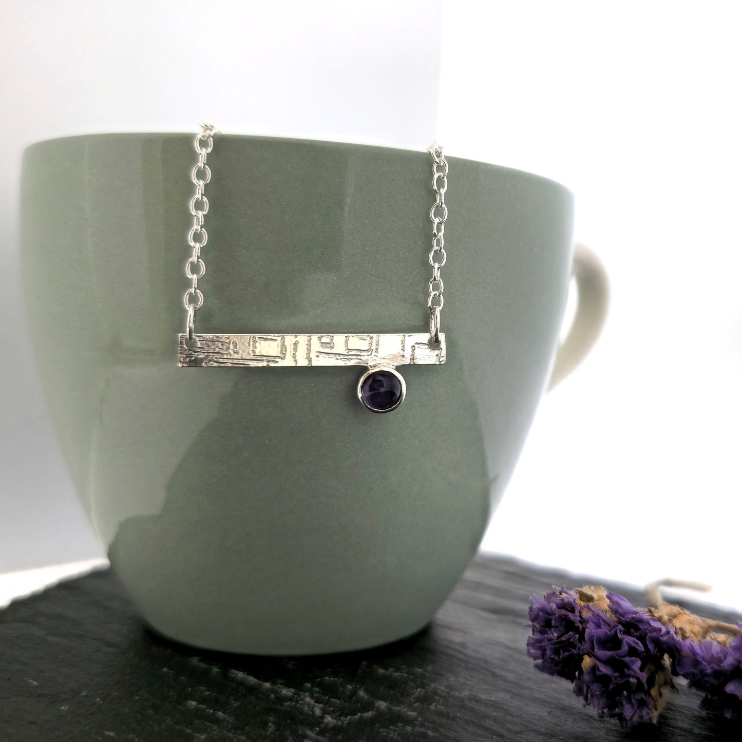 A handmade sterling silver bar necklace with a textured horizontal bar and a small round purple amethyst gemstone, displayed on a matte green ceramic cup and dried purple flowers. Mijoux Jewellery.