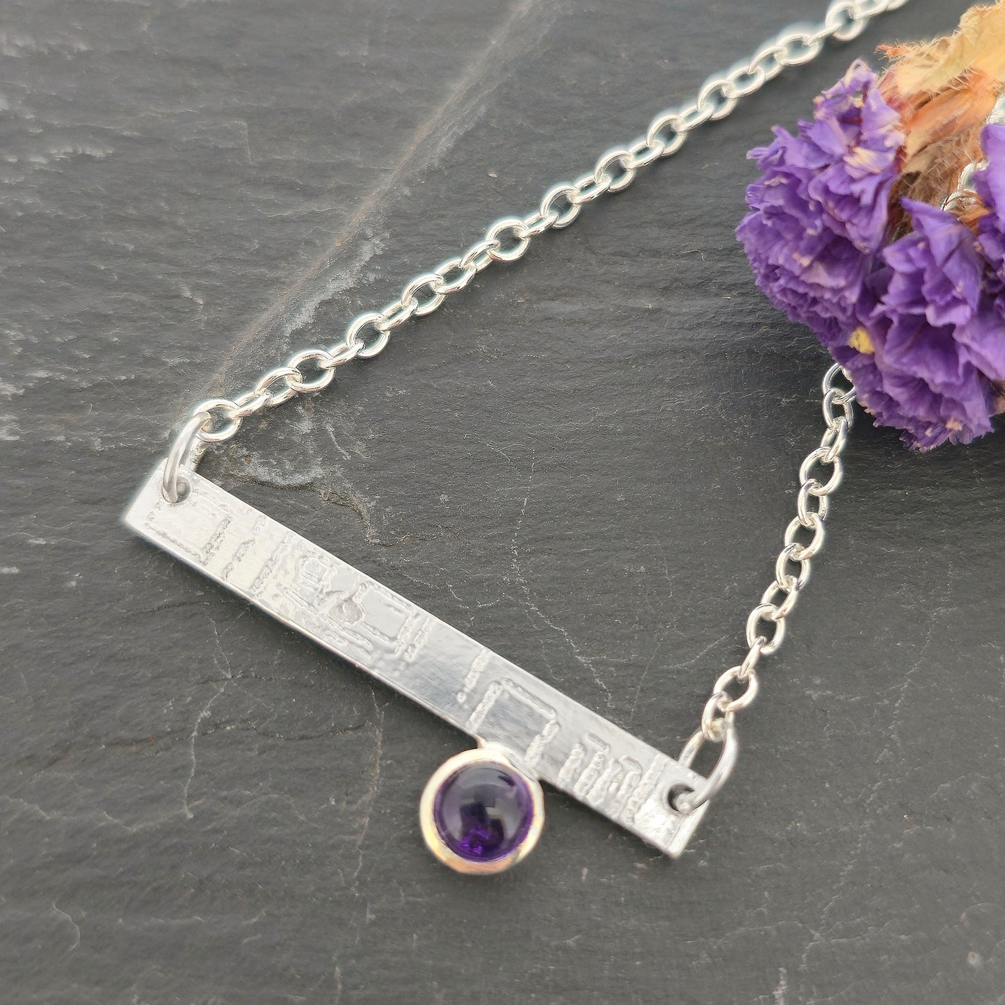 A handmade sterling silver bar necklace with engraved square pattern and a round bezel-set purple amethyst gemstone, displayed on a dark slate surface next to dried purple flowers. Mijoux Jewellery.