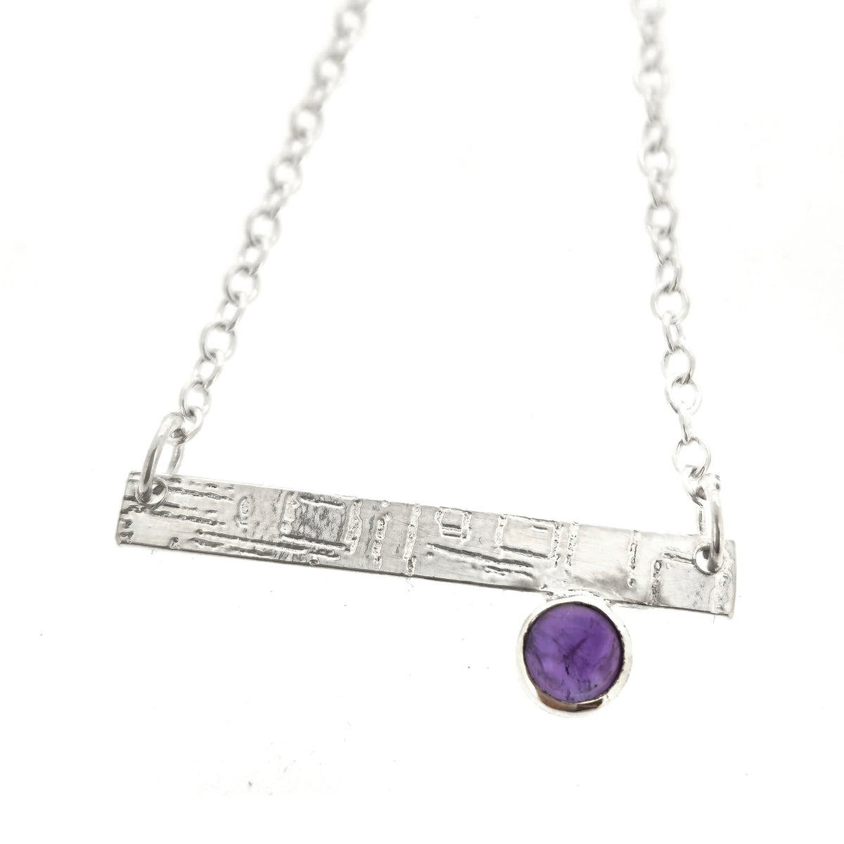 A handmade silver bar necklace with a textured horizontal bar and a small bezel-set purple amethyst stone, shown on a plain white background. Mijoux Jewellery.
