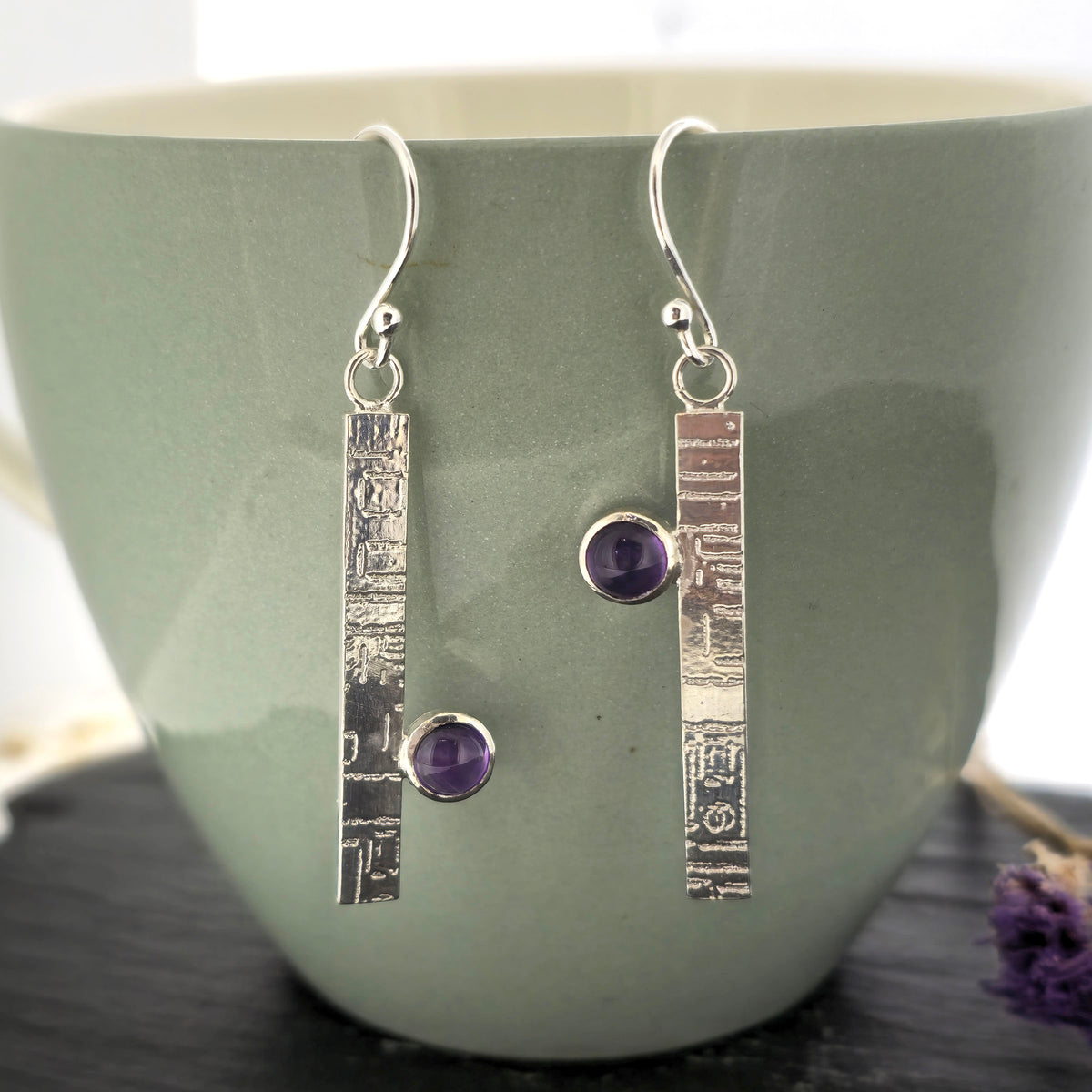 A pair of handmade silver drop earrings with textured rectangular bars and round purple amethyst gemstones, displayed against a sage green ceramic mug. Mijoux Jewellery.