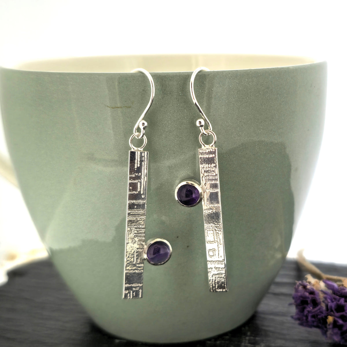 A pair of handmade silver drop earrings with textured rectangular bars and round purple amethyst gemstones, displayed against a sage green ceramic mug. Mijoux Jewellery.