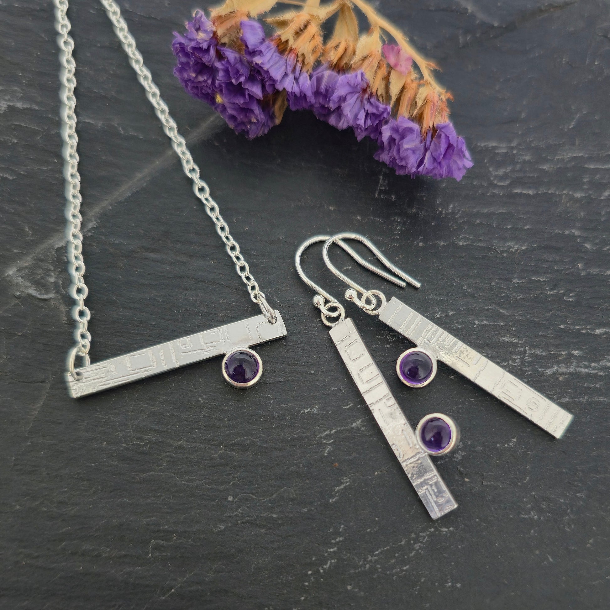 Handmade silver jewellery set including a bar necklace and matching drop earrings with small purple amethyst gemstones, displayed on a dark textured surface with dried purple flowers. Mijoux Jewellery.