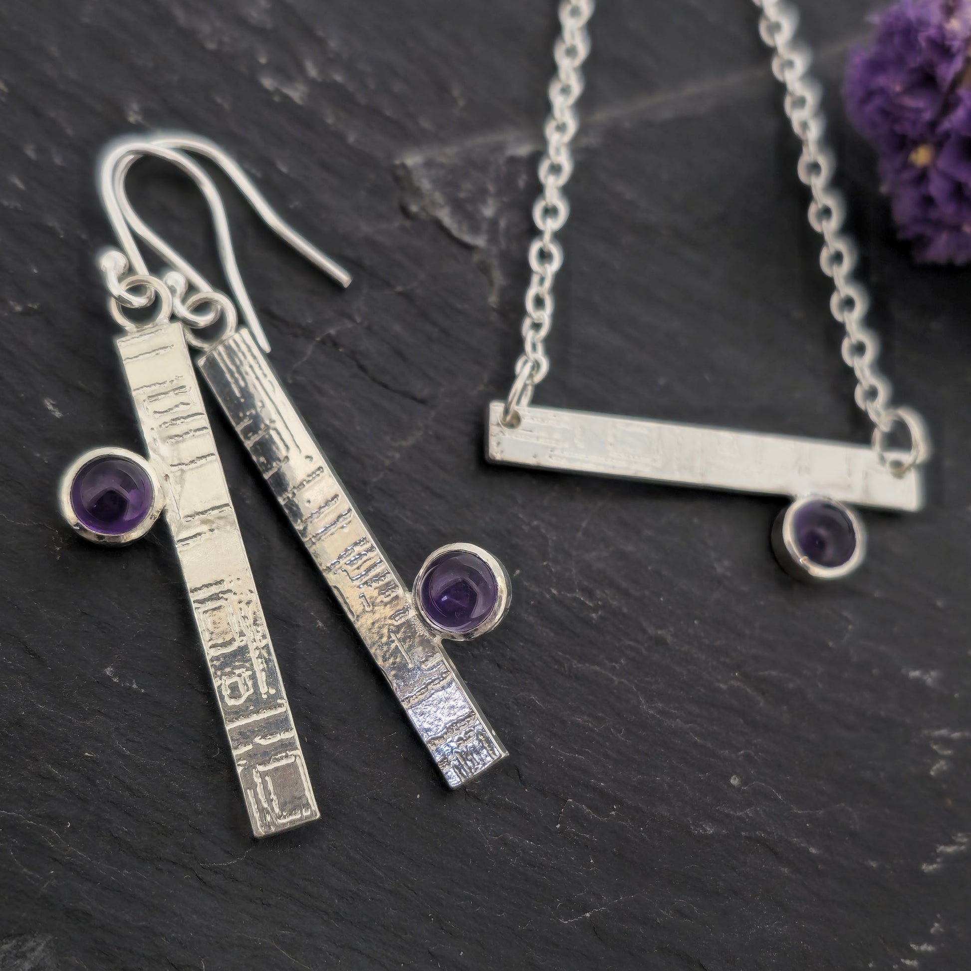 Handmade silver jewellery set including a bar necklace and matching drop earrings with small purple amethyst gemstones, displayed on a dark textured surface with dried purple flowers. Mijoux Jewellery.