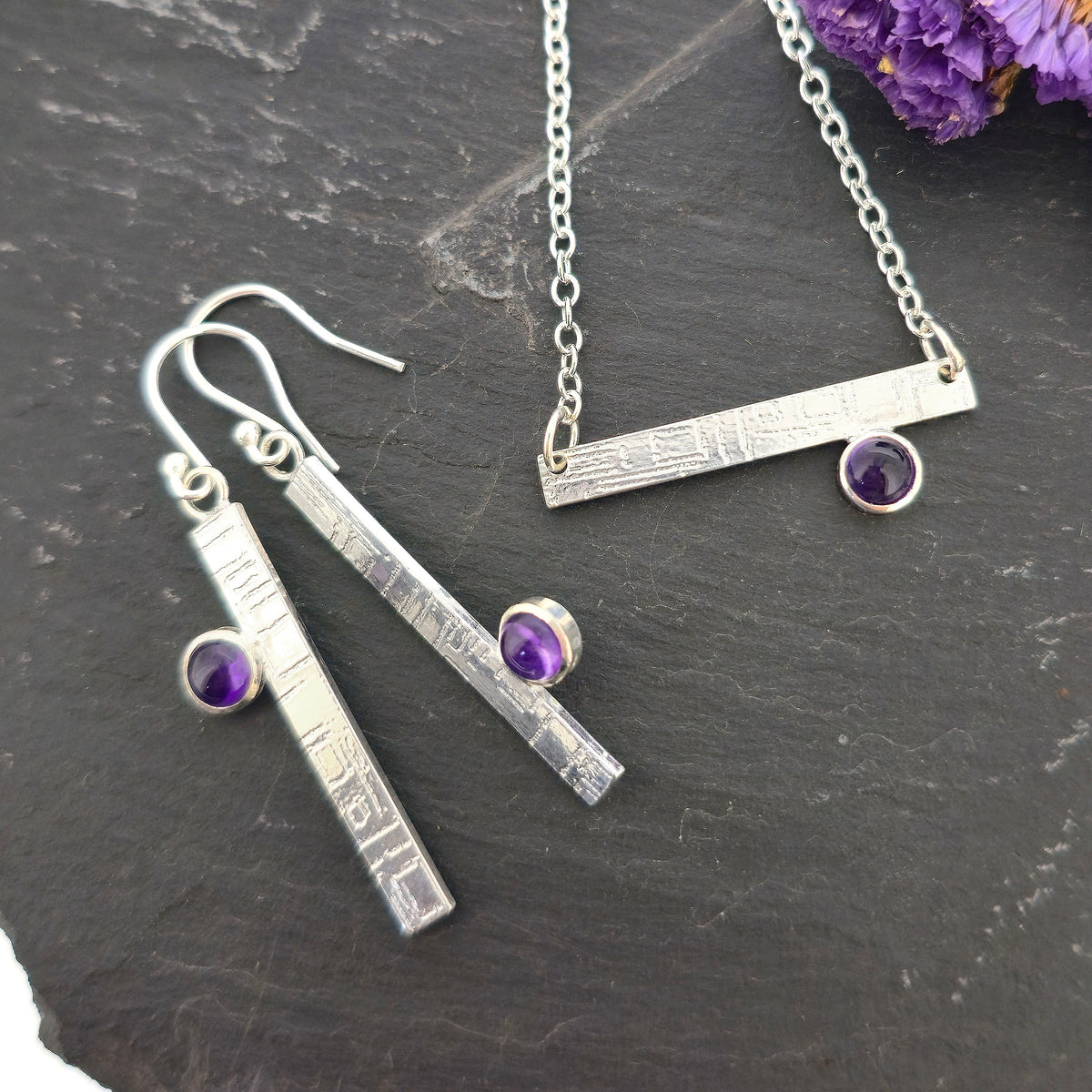 Handmade silver jewellery set including a bar necklace and matching drop earrings with small purple amethyst gemstones, displayed on a dark textured surface with dried purple flowers. Mijoux Jewellery.
