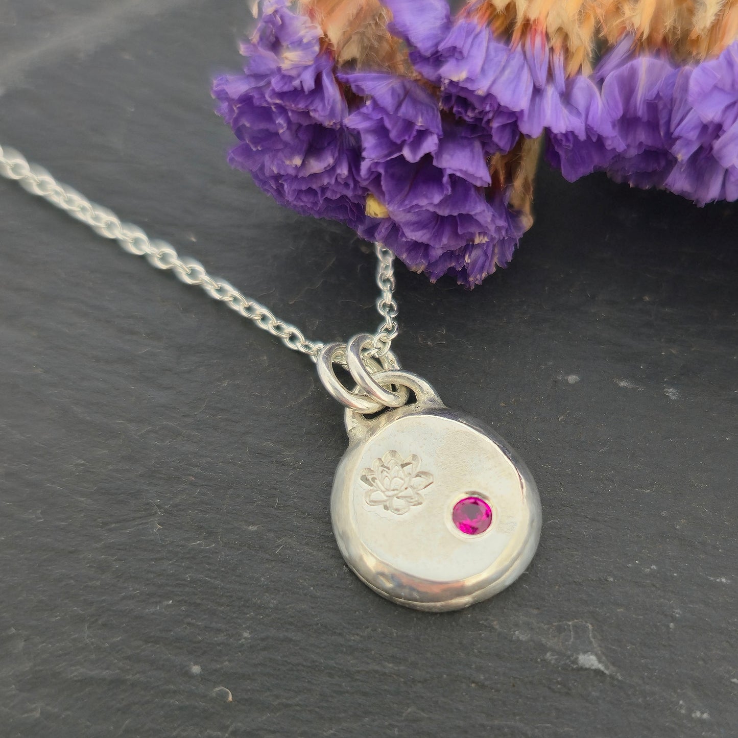 A round silver flat pebble pendant with an engraved lotus flower and flush set red ruby gemstone. On a black background with dried flowers.