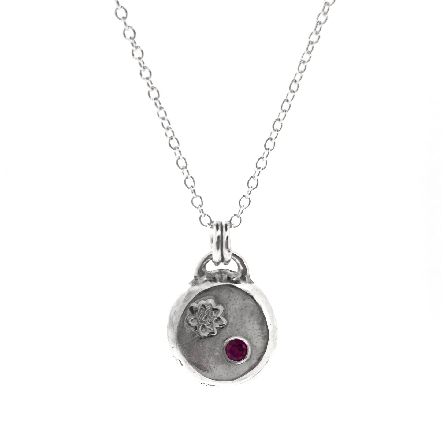 A round silver flat pebble pendant with an engraved lotus flower and flush set red ruby gemstone.