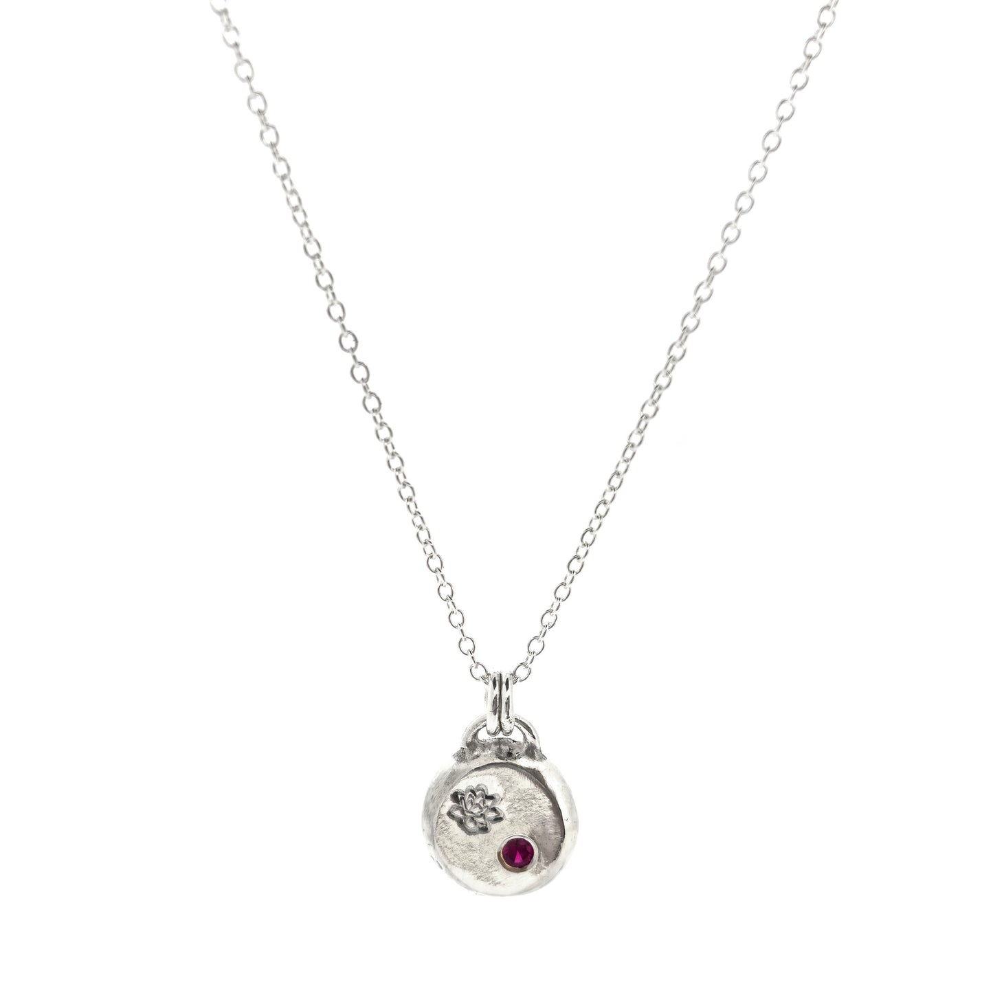 A round silver flat pebble pendant with an engraved lotus flower and flush set red ruby gemstone.