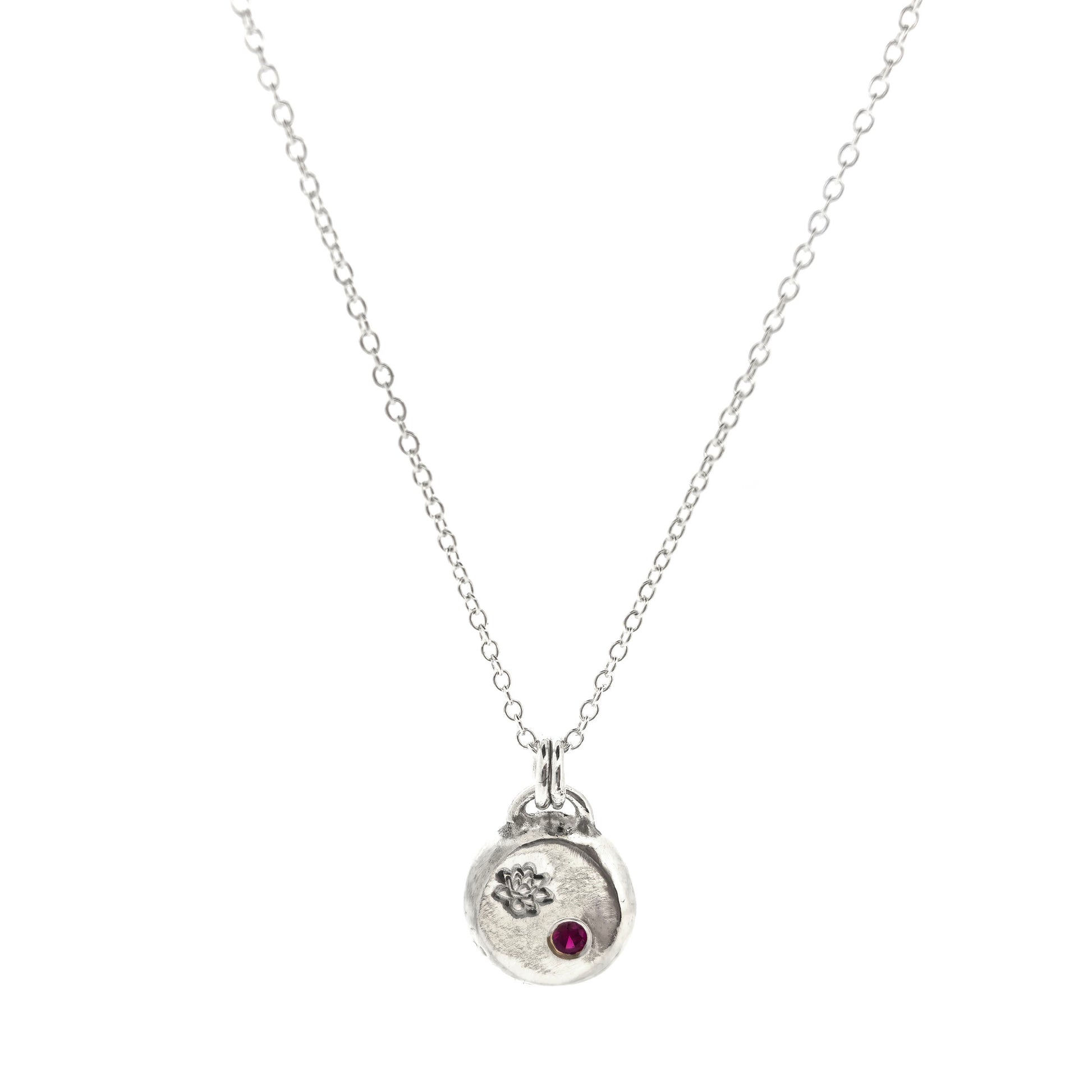 A round silver flat pebble pendant with an engraved lotus flower and flush set red ruby gemstone.