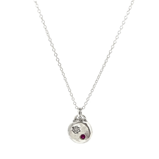A round silver flat pebble pendant with an engraved lotus flower and flush set red ruby gemstone.