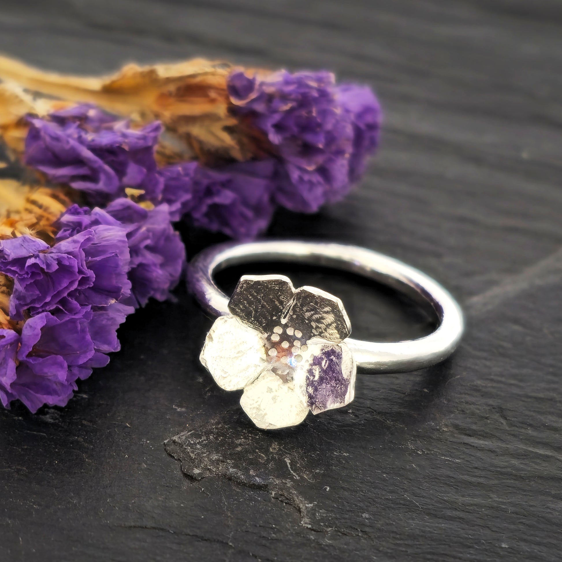 A handmade silver 5 petal flower stacking ring with textured petals, displayed on a dark slate surface next to dried purple flowers. Mijoux Jewellery.