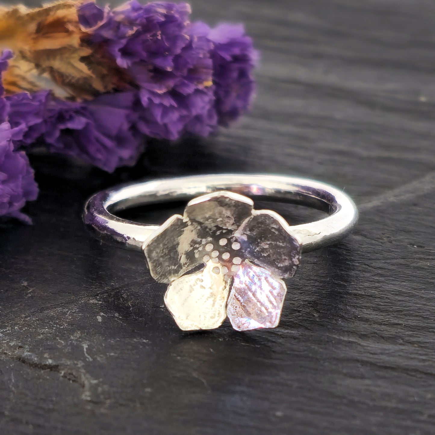 A handmade silver 5 petal flower stacking ring with textured petals, displayed on a dark slate surface next to dried purple flowers. Mijoux Jewellery.