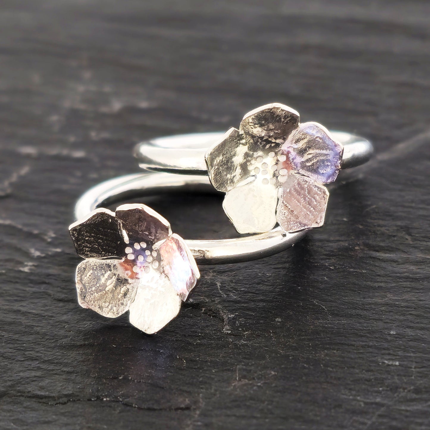 A pair of handmade silver 5 petal flower rings with textured petals, displayed on a dark slate surface. Mijoux Jewellery.