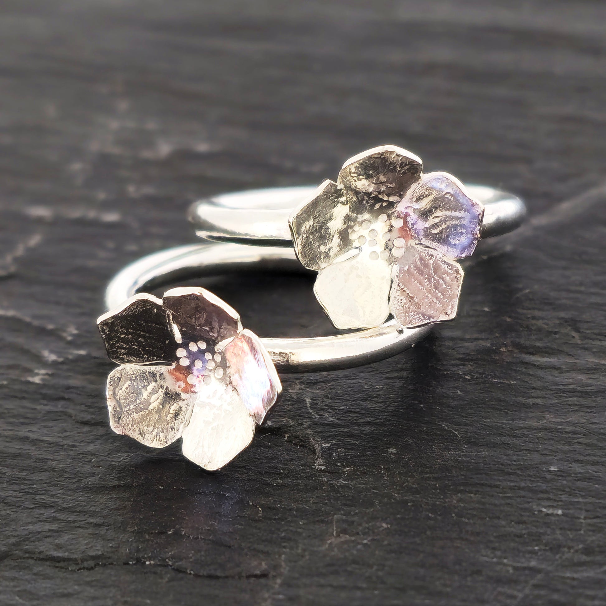 A pair of handmade silver 5 petal flower rings with textured petals, displayed on a dark slate surface. Mijoux Jewellery.