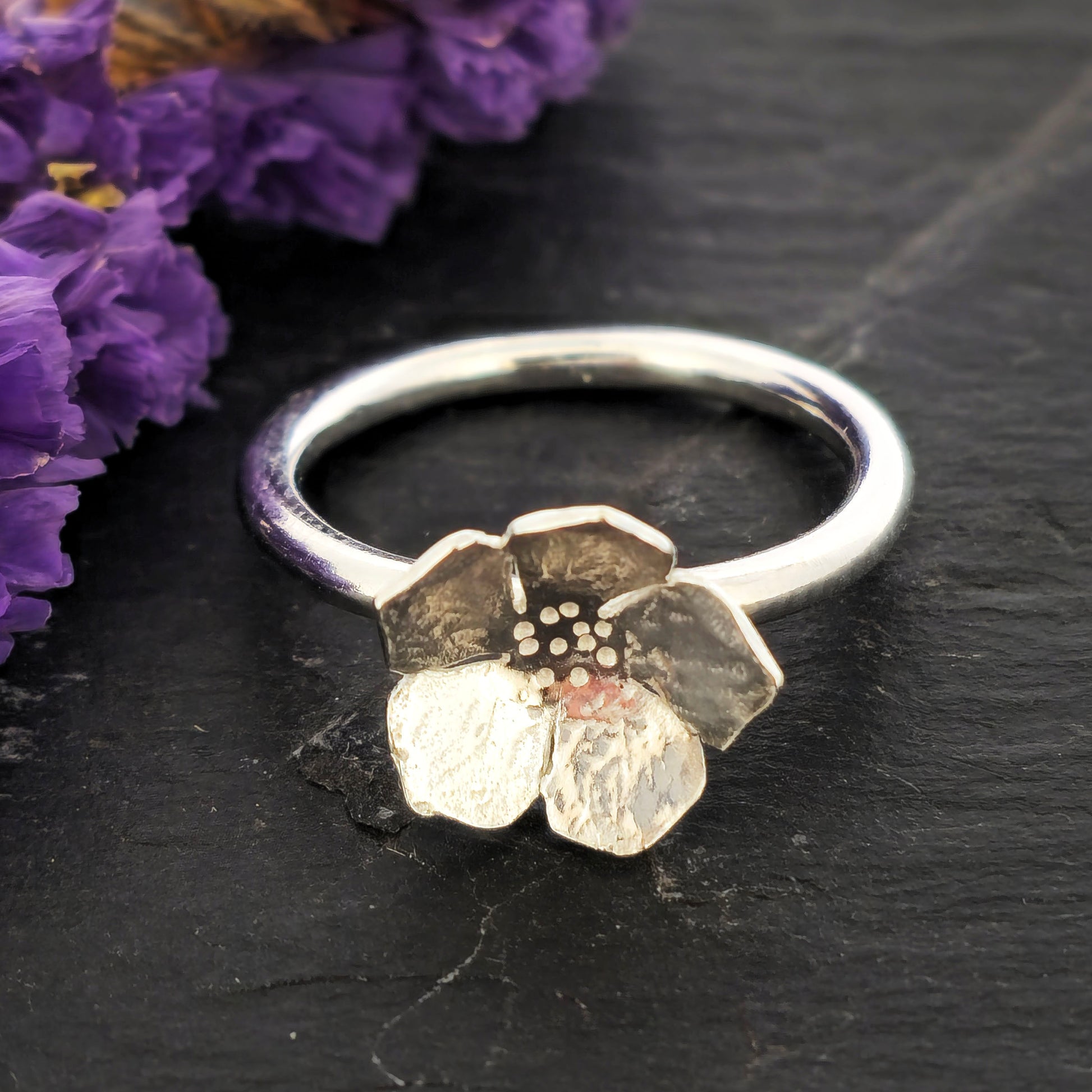 A handmade silver 5 petal flower stacking ring with textured petals, displayed on a dark slate surface next to dried purple flowers. Mijoux Jewellery.