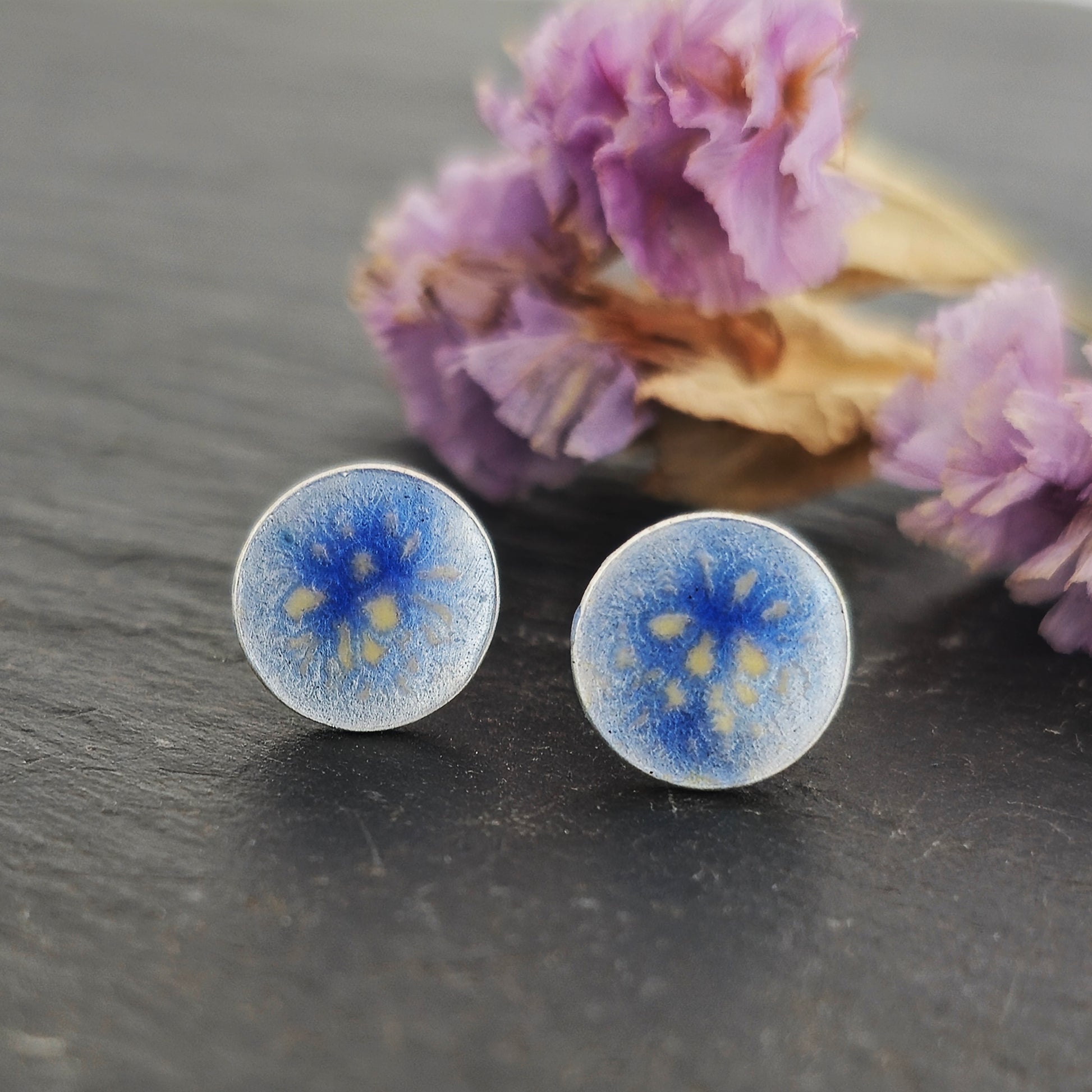 Round silver stud earrings with a blue and white enamel speckled pattern. Shown on slate with flowers.