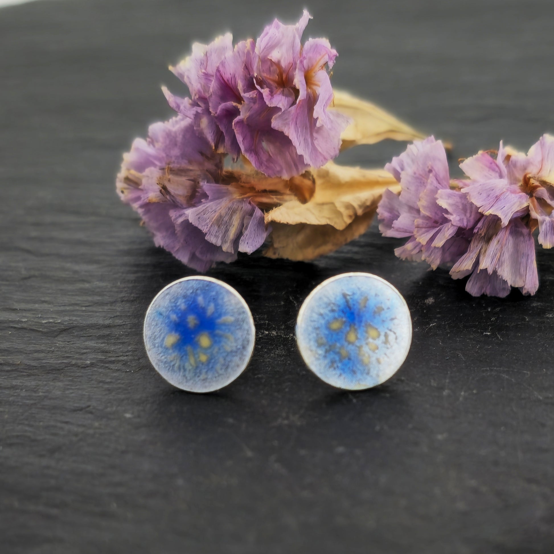 Round silver stud earrings with a blue and white enamel speckled pattern. Shown on slate with flowers.