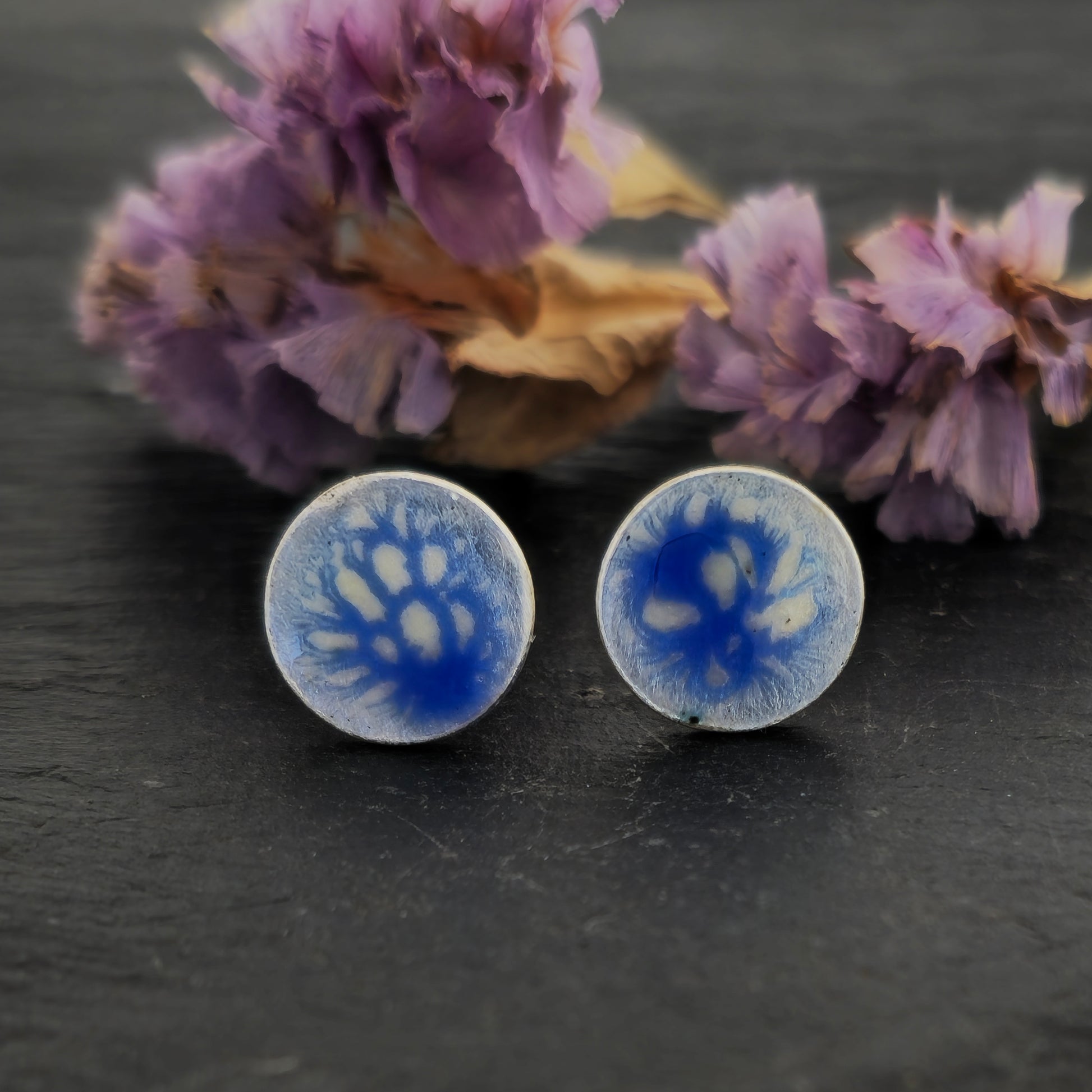 Round silver stud earrings with a blue and white enamel speckled pattern. Shown on slate with flowers.