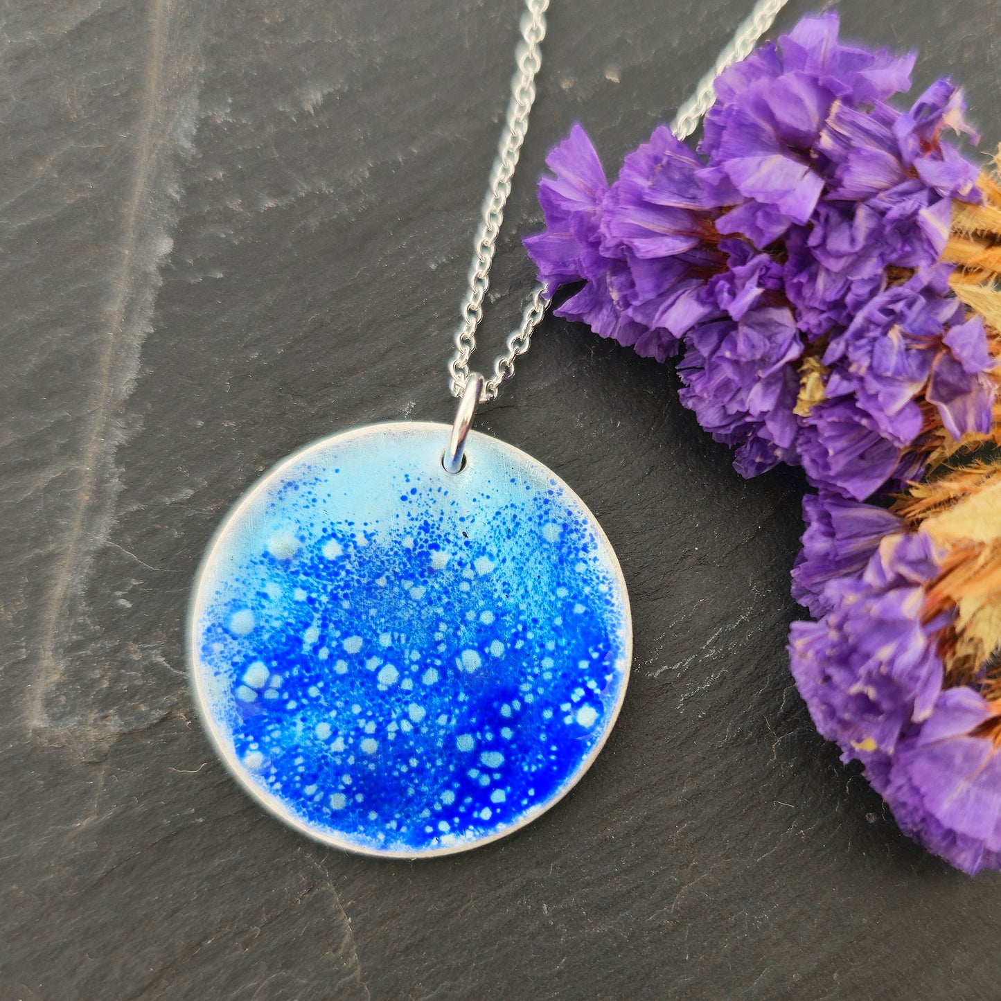 A silver pendant necklace with a round disc pendant featuring a blue and white cosmic glitter pattern, displayed on a dark slate surface next to purple dried flowers. Mijoux Jewellery.