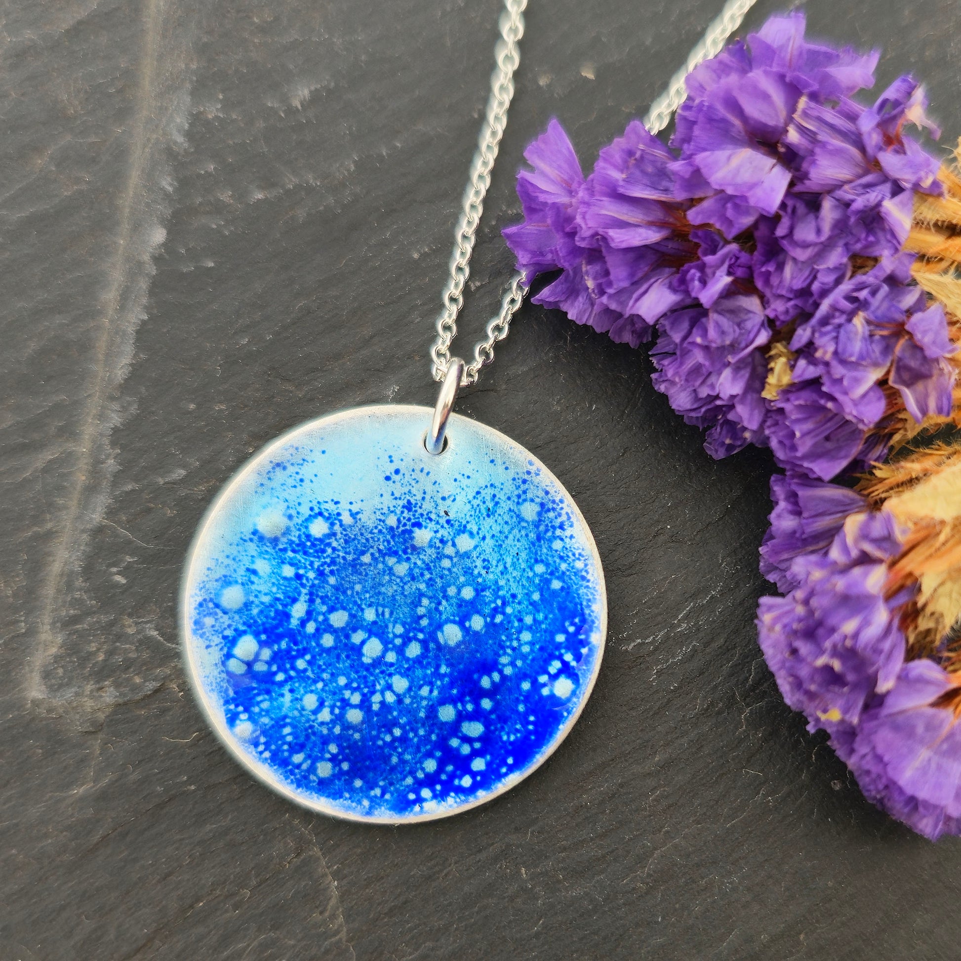 A silver pendant necklace with a round disc pendant featuring a blue and white cosmic glitter pattern, displayed on a dark slate surface next to purple dried flowers. Mijoux Jewellery.