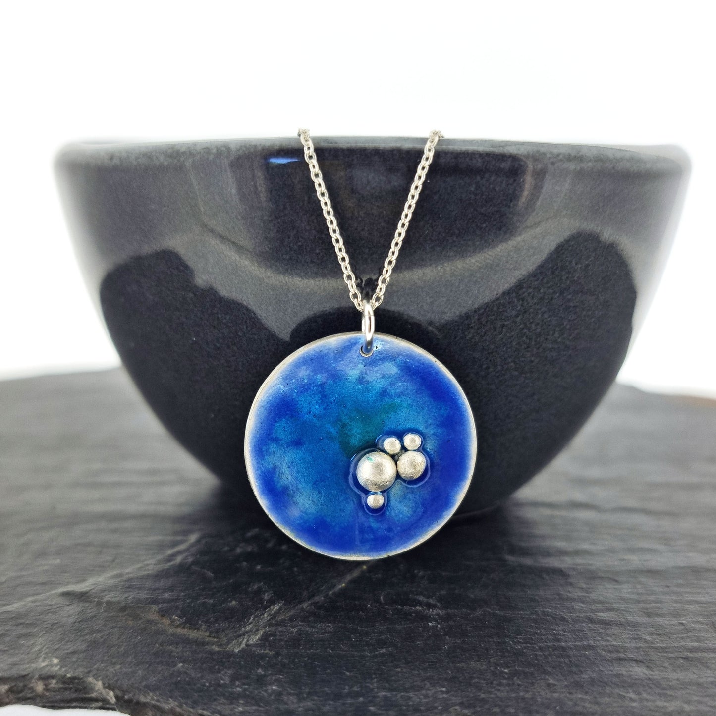 A round silver pendant with blue, turquoise and green enamel and 5 silver balls in a cluster in the bottom right hand of the circle. On a silver chain. Pictured on a bowl.