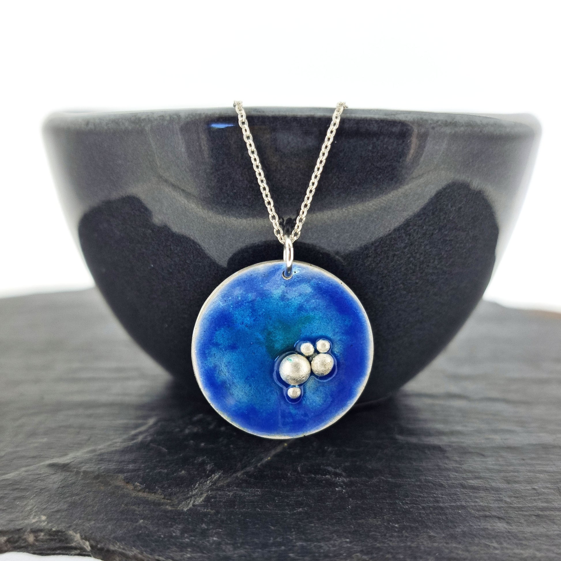 A round silver pendant with blue, turquoise and green enamel and 5 silver balls in a cluster in the bottom right hand of the circle. On a silver chain. Pictured on a bowl.