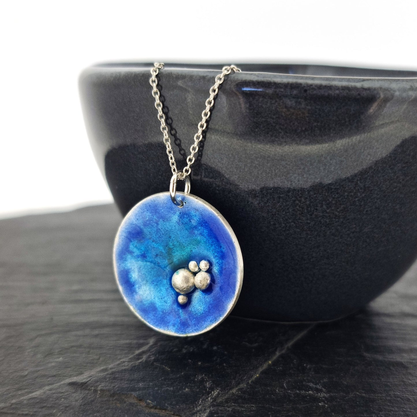 A round silver pendant with blue, turquoise and green enamel and 5 silver balls in a cluster in the bottom right hand of the circle. On a silver chain. Pictured on a bowl.