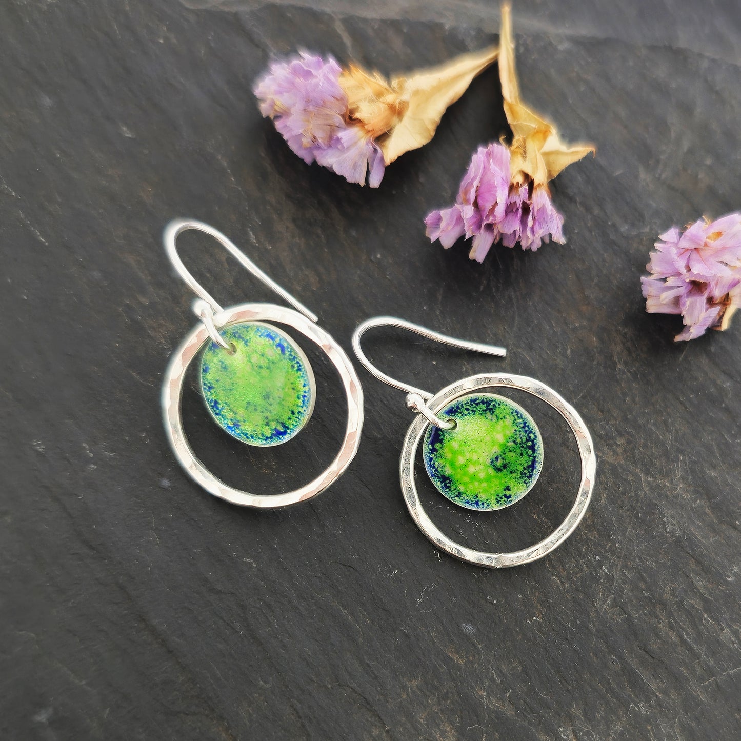 Silver drop earrings consisting of a central disc with a pattern of green and blue enamel surrounded by a silver circle with a hammered finish. On silver ear hooks. Shown on slate with flowers.