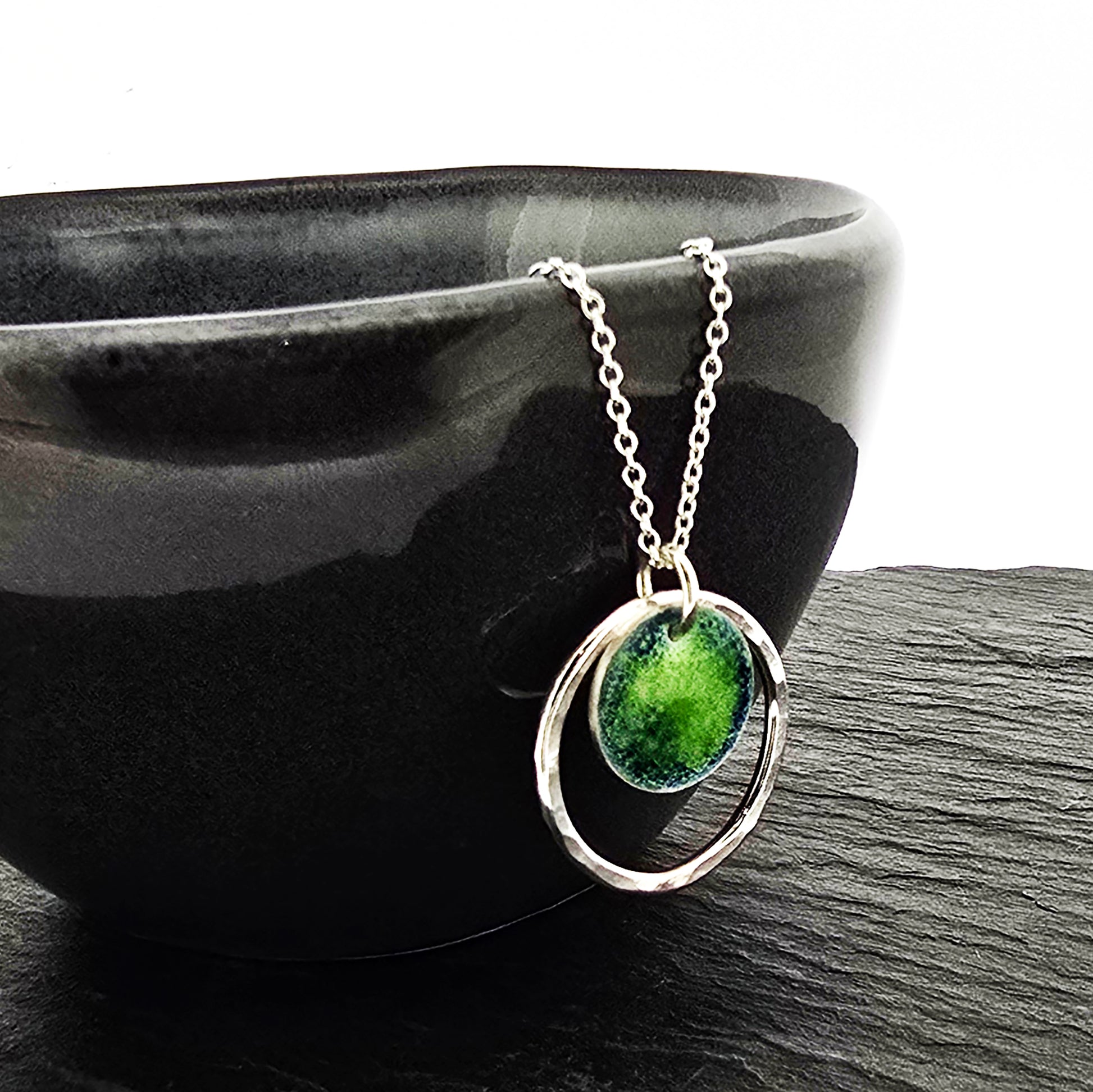 A silver pendant necklace featuring a silver disc with a pattern of green and blue enamel surrounded by a silver circle with a hammered finish. On a silver chain. Shown on a black bowl.