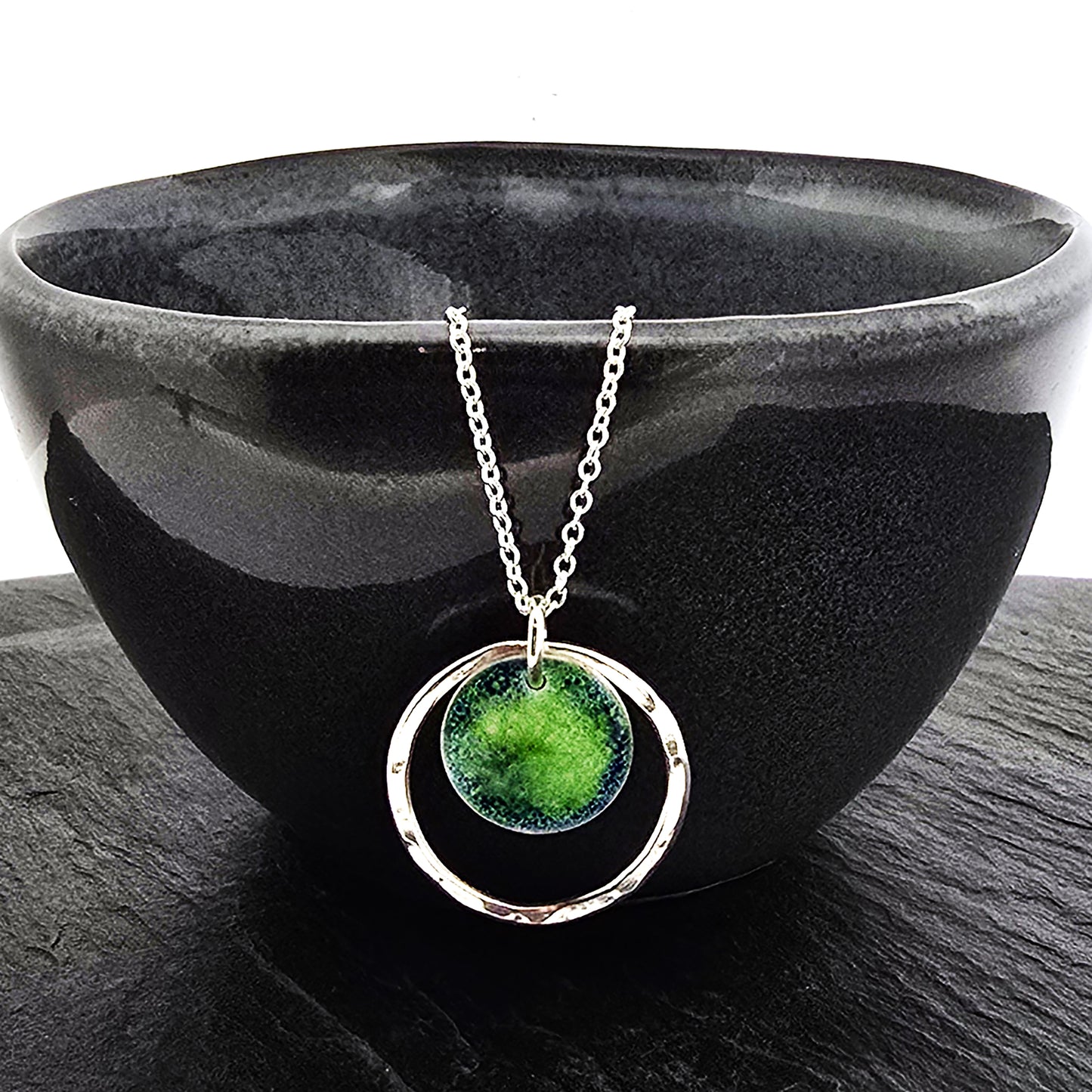 A silver pendant necklace featuring a silver disc with a pattern of green and blue enamel surrounded by a silver circle with a hammered finish. On a silver chain. Shown on a black bowl.