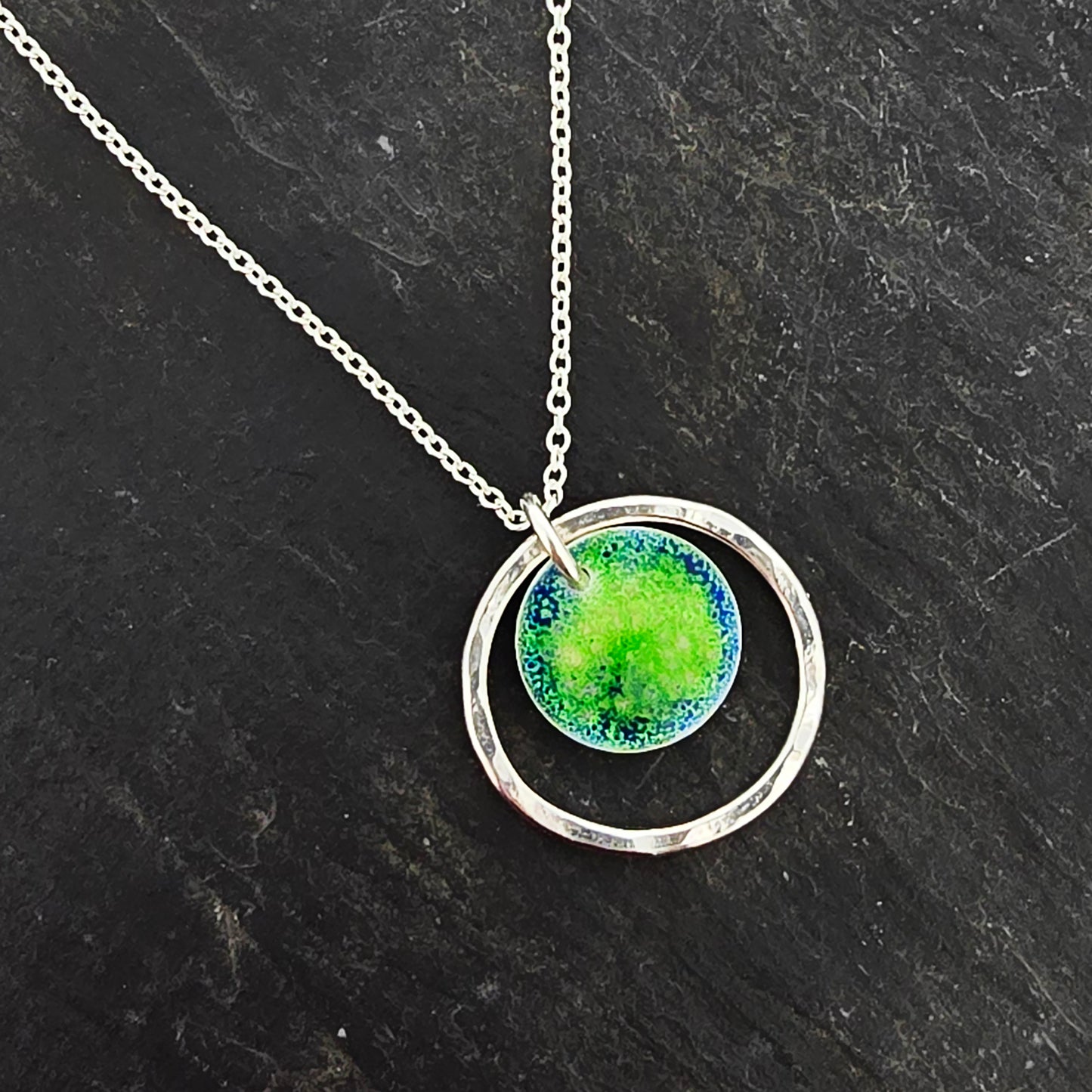 A silver pendant necklace featuring a silver disc with a pattern of green and blue enamel surrounded by a silver circle with a hammered finish. On a silver chain. Shown on a black slate.