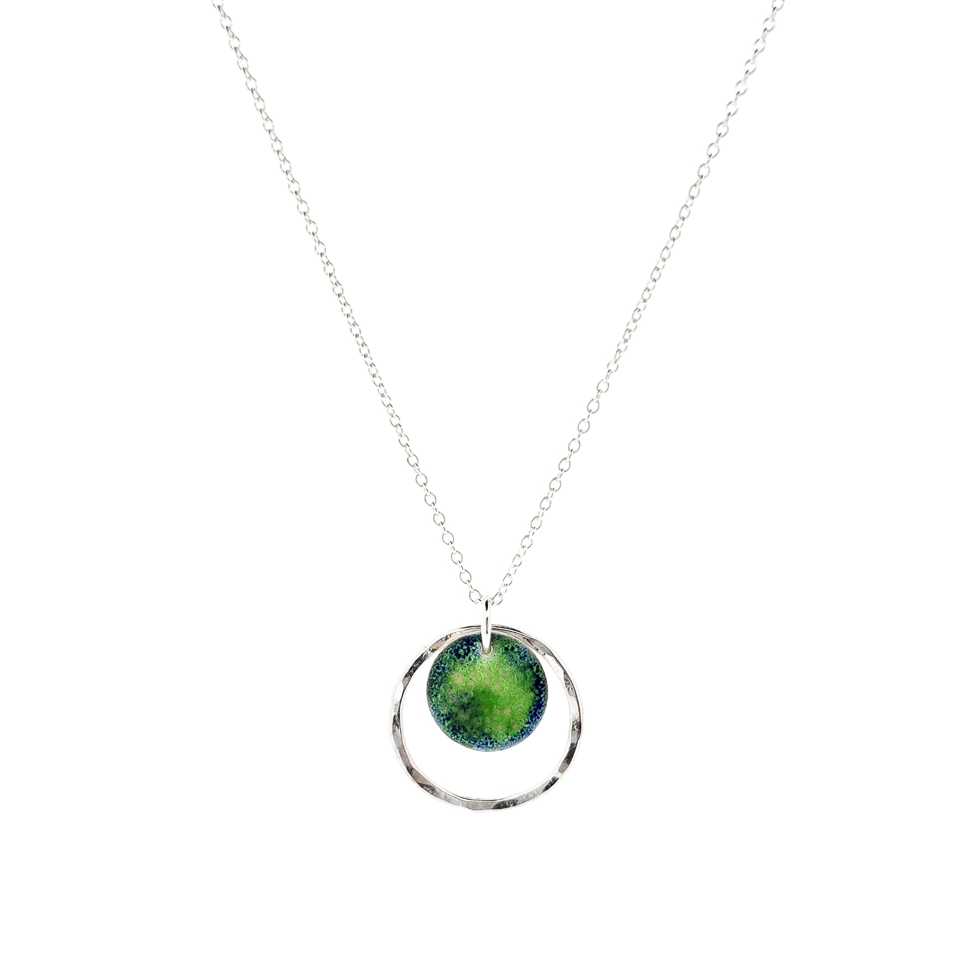 A silver pendant necklace featuring a silver disc with a pattern of green and blue enamel surrounded by a silver circle with a hammered finish. On a silver chain. 