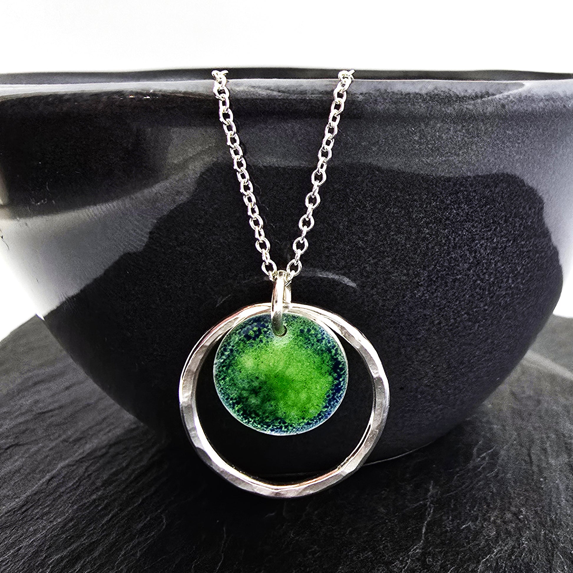 A silver pendant necklace featuring a silver disc with a pattern of green and blue enamel surrounded by a silver circle with a hammered finish. On a silver chain. Shown on a black bowl.