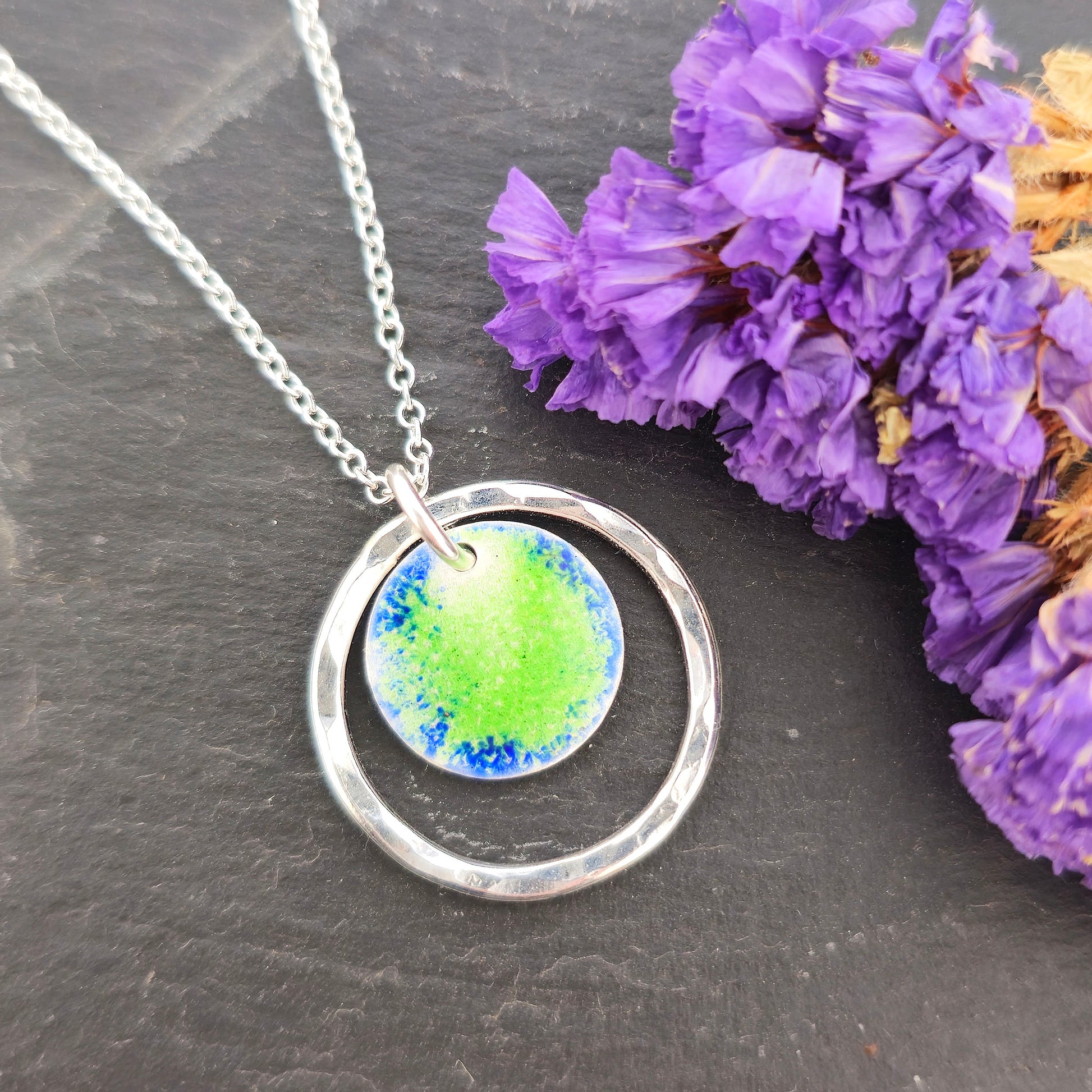 A silver pendant necklace featuring a silver disc with a pattern of green and blue enamel surrounded by a silver circle with a hammered finish. On a silver chain. Shown on a black background with purple flowers. Mijoux Jewellery.