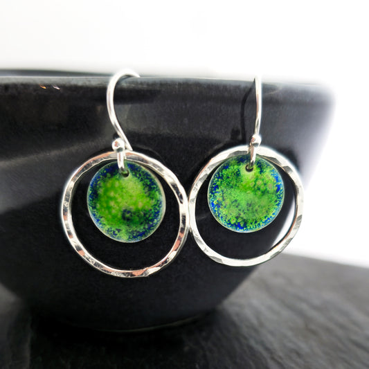 Silver drop earrings consisting of a central disc with a pattern of green and blue enamel surrounded by a silver circle with a hammered finish. On silver ear hooks. Pictured on a black bowl.