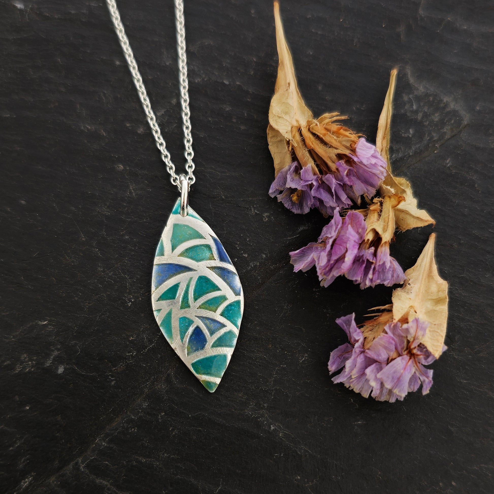 A marquise shaped silver pendant with geometric cells of blue, green and turquoise enamel. On a silver chain. Large. Pictured on slate with flowers.