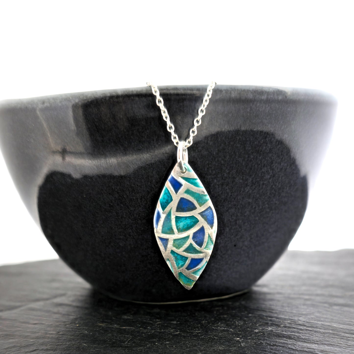 A marquise shaped silver pendant with geometric cells of blue, green and turquoise enamel. On a silver chain. Large. Pictured on a bowl.