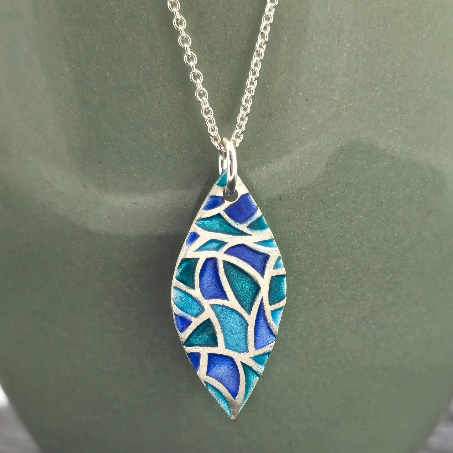 A marquise shaped silver pendant with geometric cells of blue, green and turquoise enamel. On a silver chain. Large. On a green background.