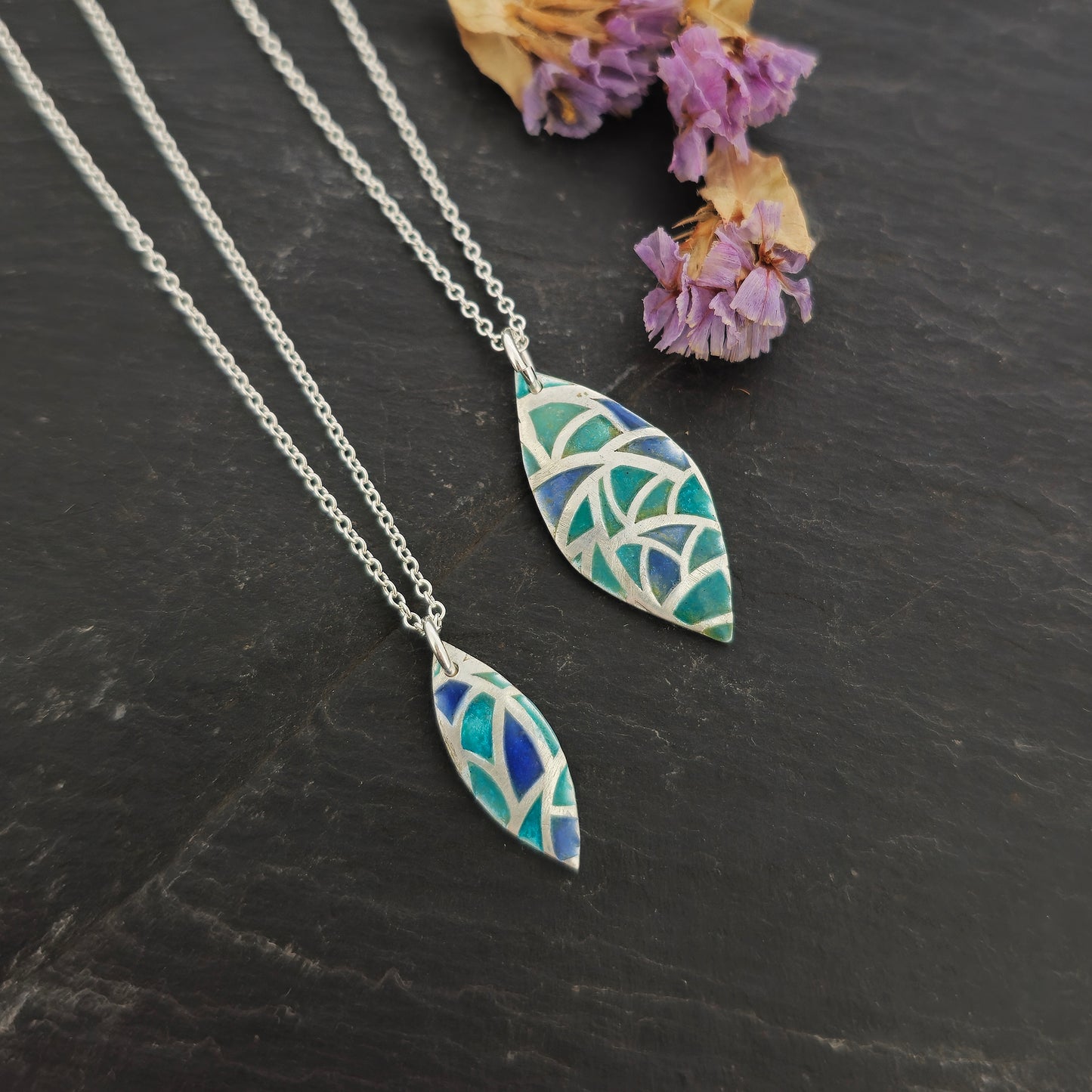 Two marquise shaped silver pendants with geometric cells of blue, green and turquoise enamel. On silver chains. Pictured on slate with flowers.