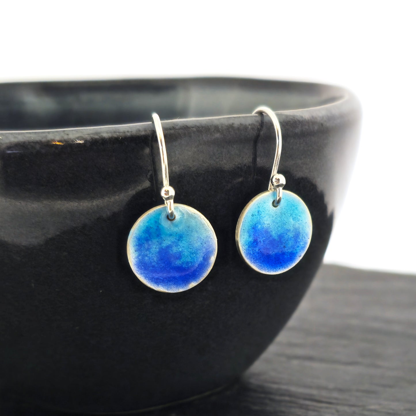 Silver round drop earrings with a mix of dark and light blue enamel. Pictured on a black bowl.